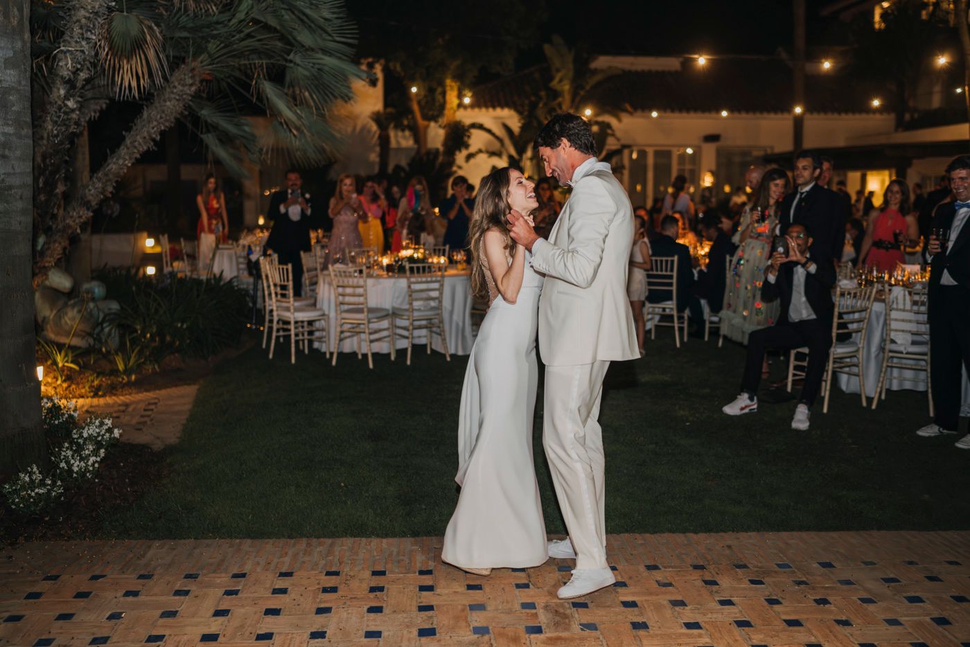 A couple shares their first dance at an outdoor evening wedding reception with string lights and palm trees.