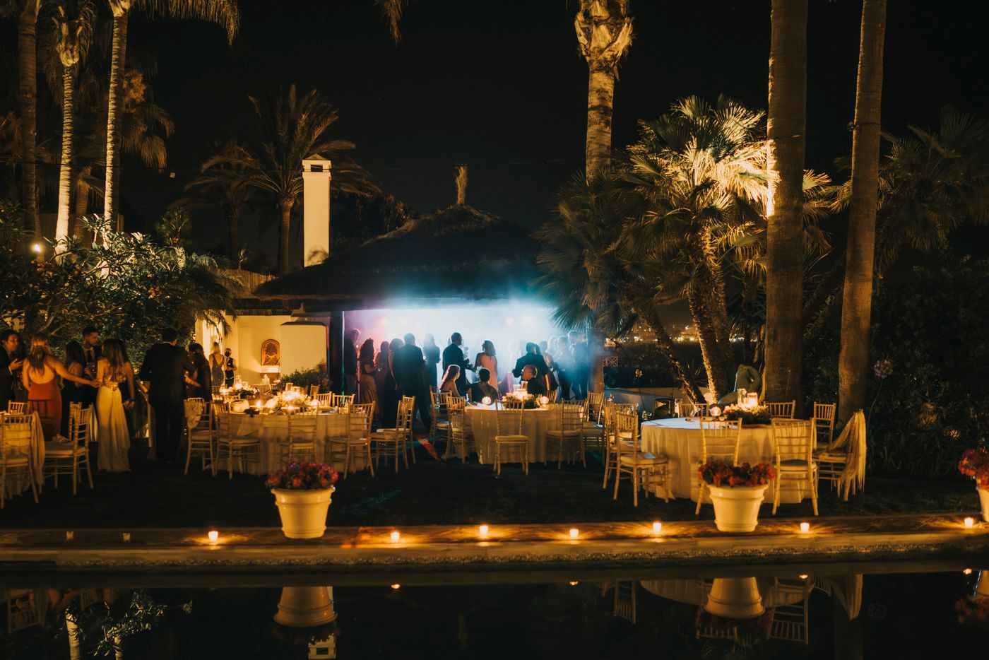 Outdoor evening party setup with glowing lights, poolside tent and palm trees creating a romantic ambiance.