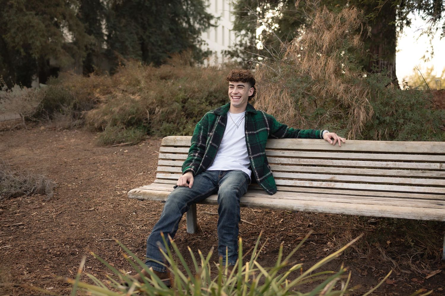 Senior student graduation photo of boy in green plaid shirt sitting on a bench in the UC Davis arboretum with an arm draped across the back