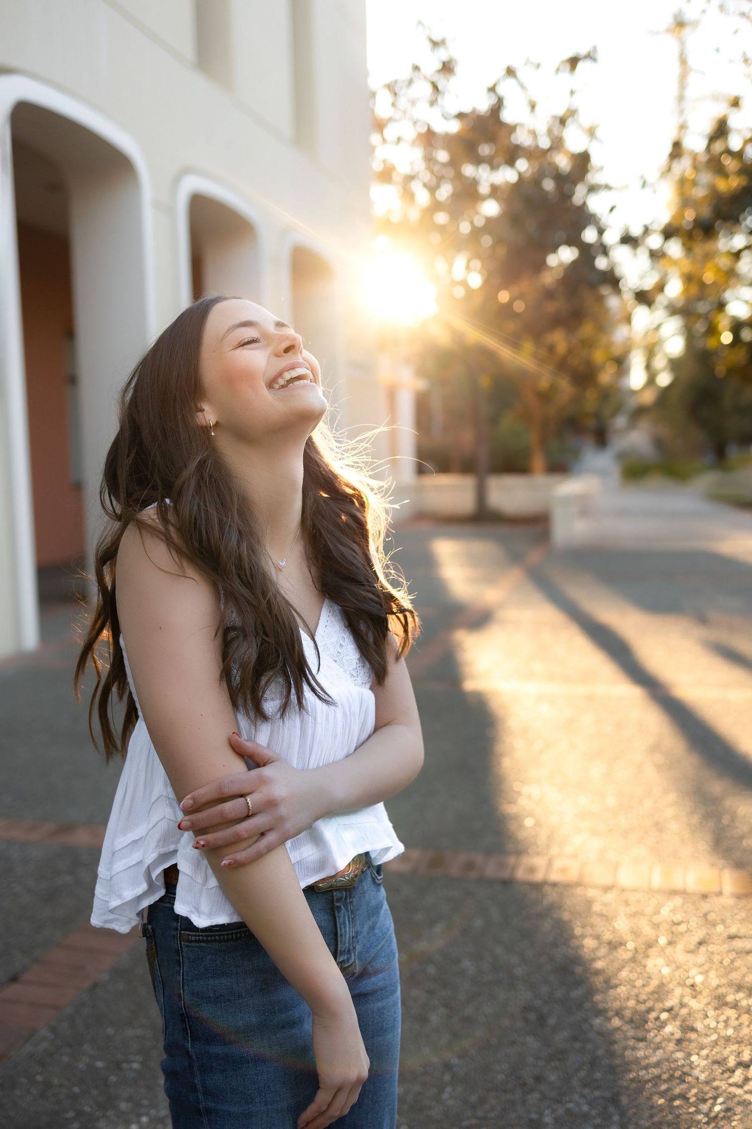 High school girl in white top with long brown hair throwing her head back and laughing with sunbeams peeking around the building behind