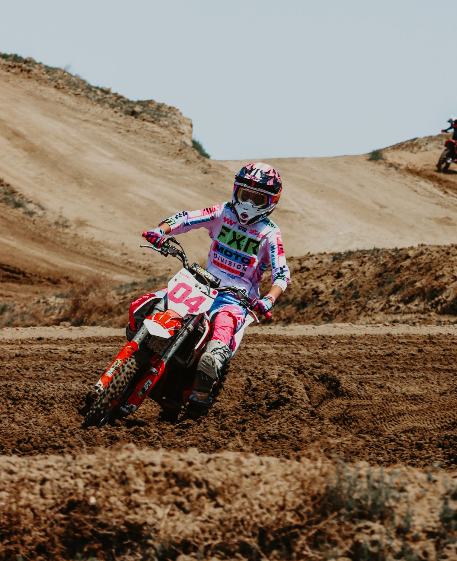 A motocross rider in pink gear races through a dirt track on a Honda motorcycle against a dusty desert backdrop.