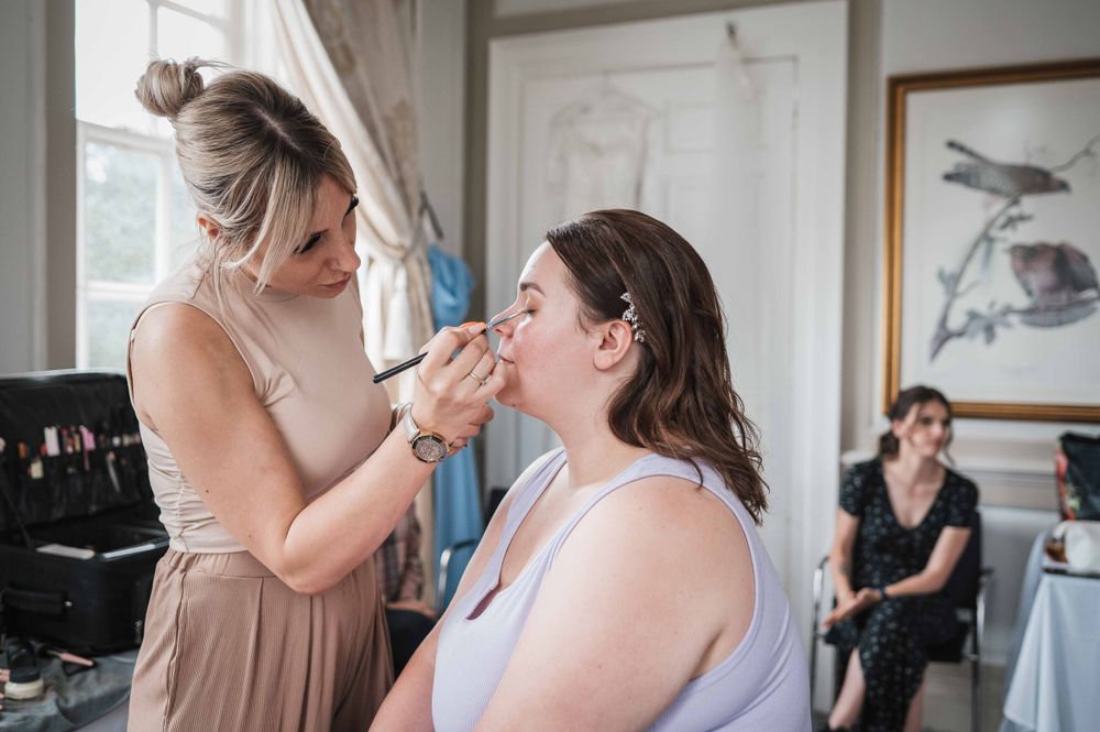 Makeup artist applies finishing touches to someone before an event while another person looks on in the background.