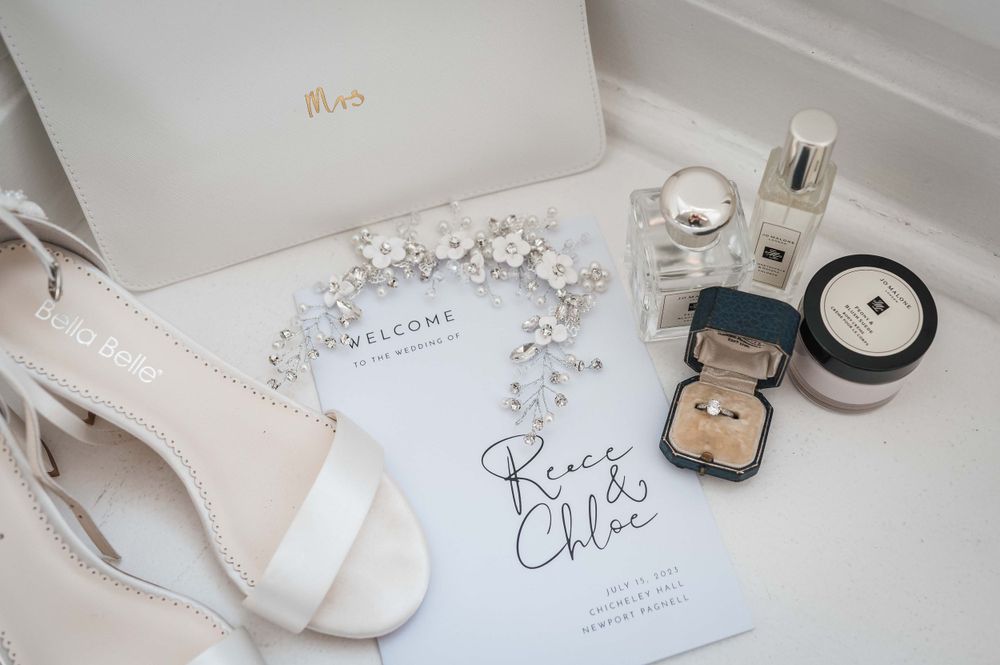 Wedding accessories including bridal shoes, invitation, jewelry box, and perfume arranged on white surface.