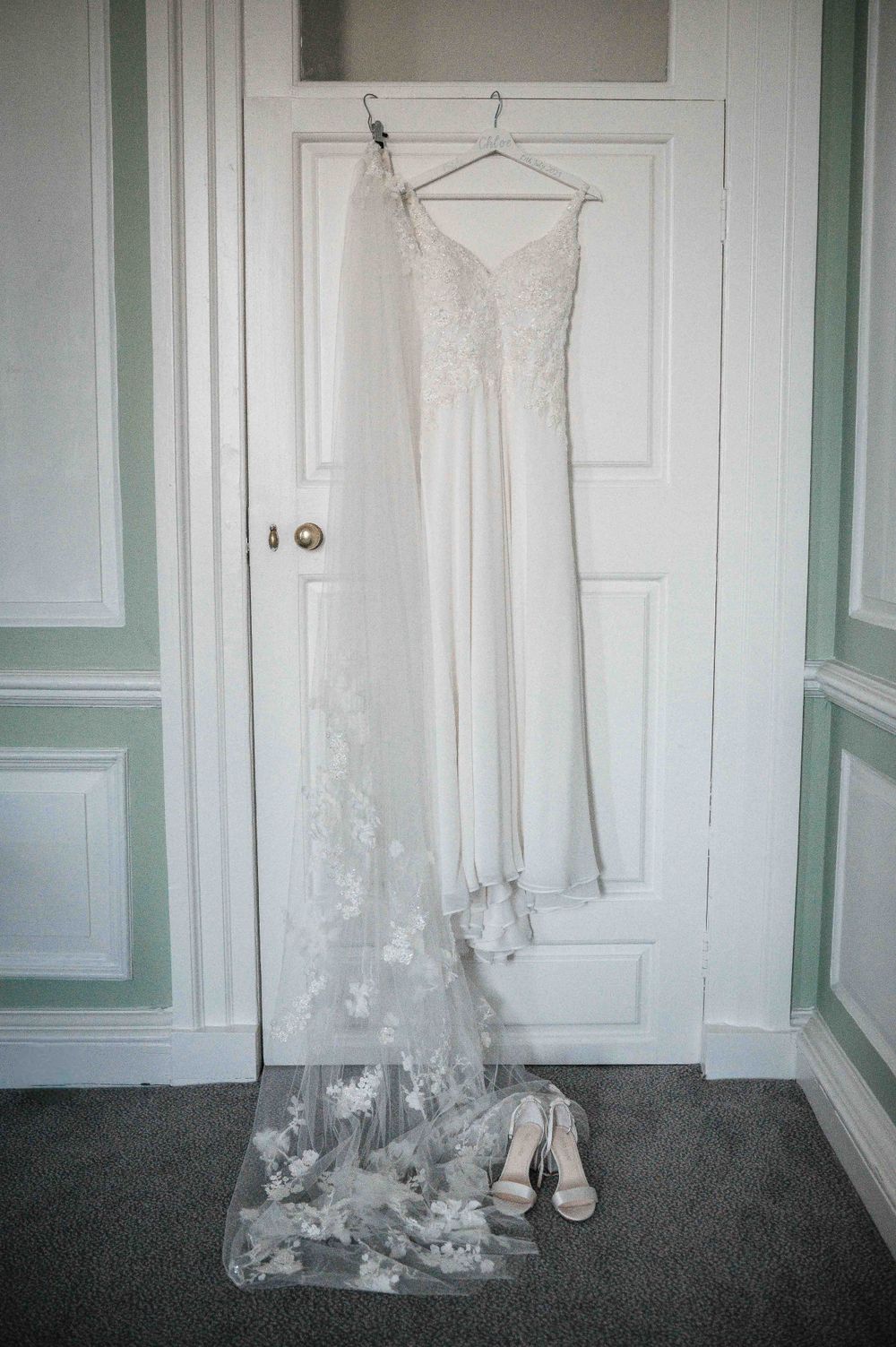 An elegant white wedding dress with lace details and a long train hangs on a door with matching white heels beneath it.