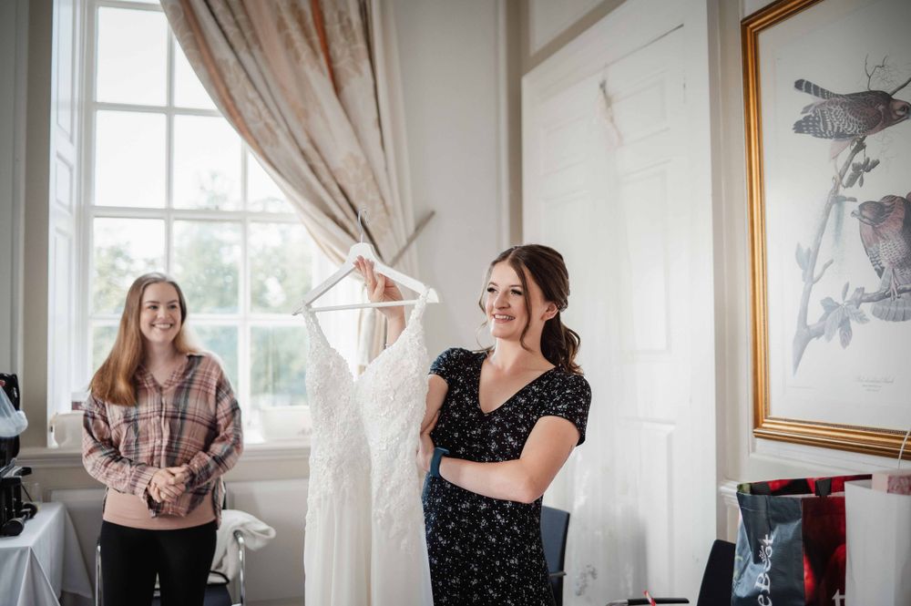Two women stand in a bright room while admiring a white wedding dress hanging on display.
