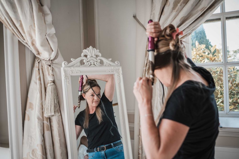 A reflection in an ornate white mirror shows someone styling their hair in a bright sunlit room.