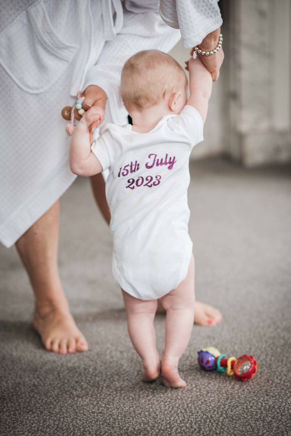 Baby in white onesie with 15th July 2023 text standing while holding onto someone's legs.