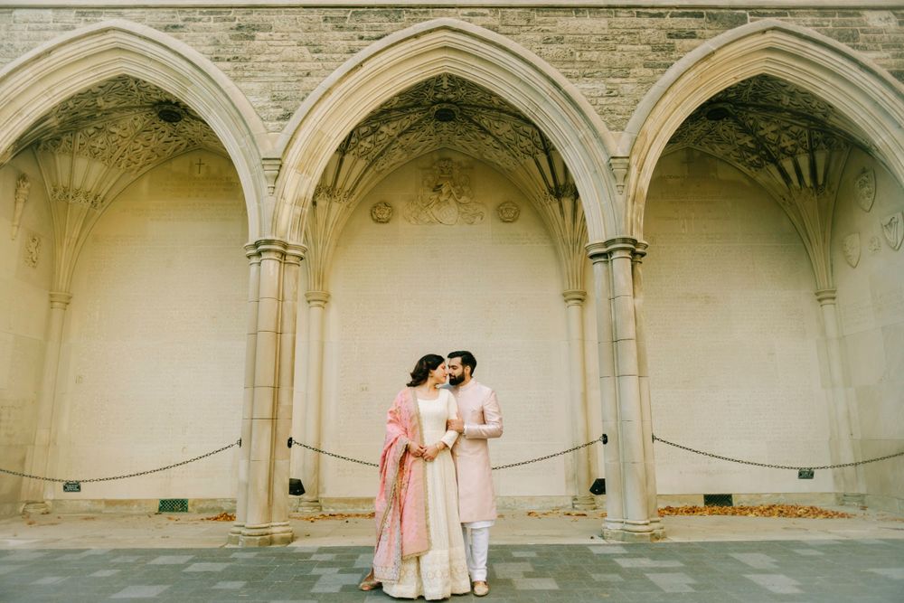 A couple in traditional pink and white attire poses under ornate stone archways in a vintage-style photograph.