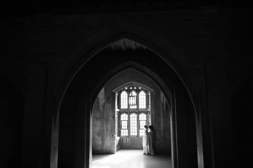 Black and white view through a gothic archway showing a tall window at the end of a dark stone corridor with a figure silhouetted.