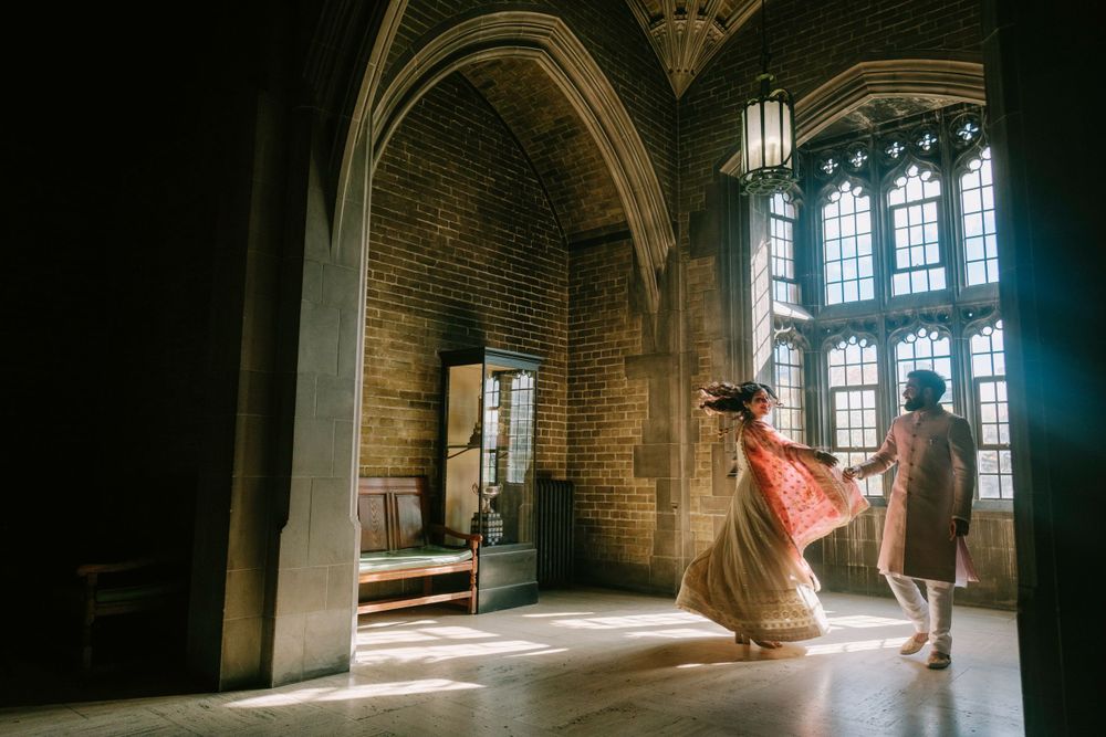 Two figures dance in a dramatic gothic hallway with arched windows and stone walls illuminated by natural light.