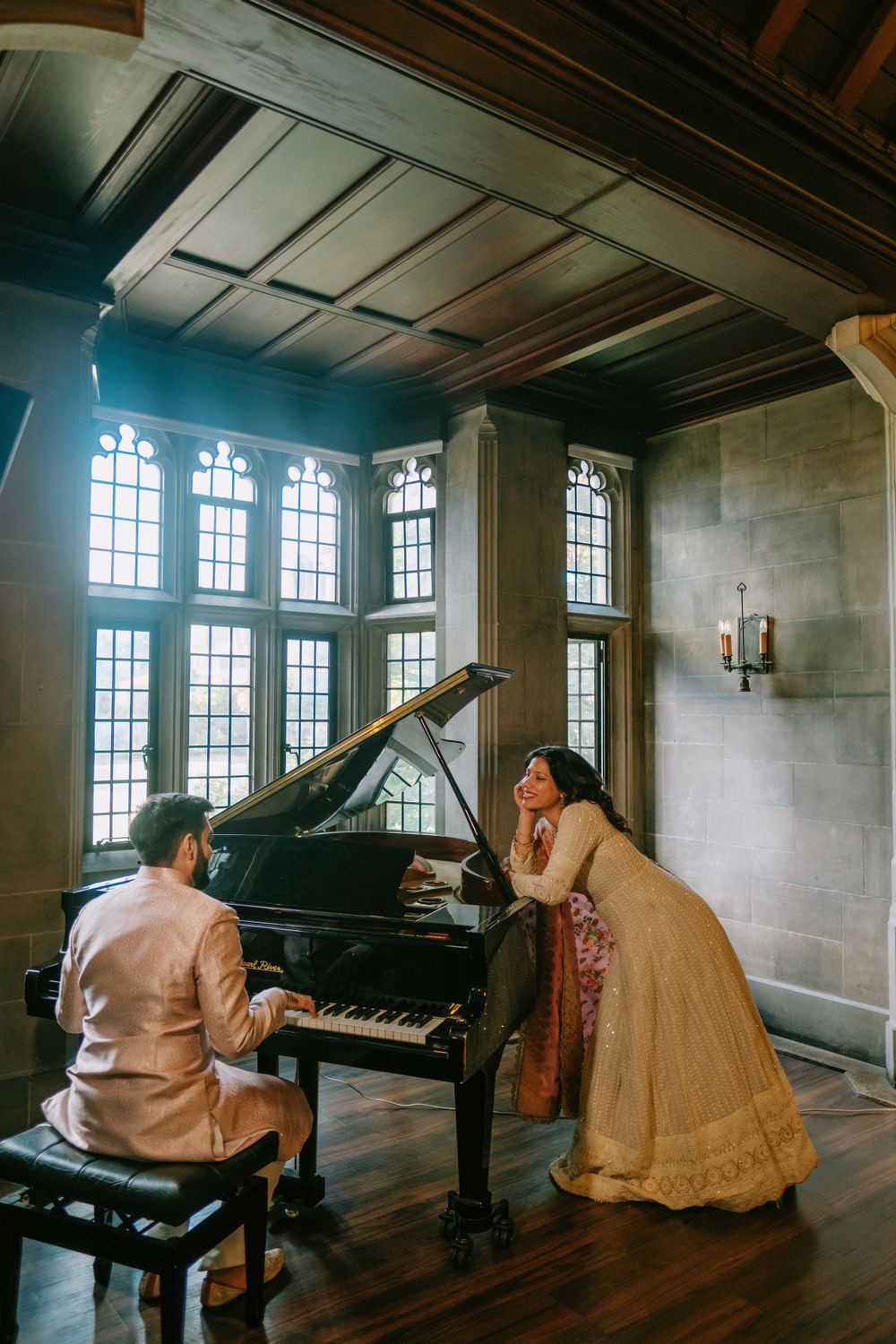 Musicians perform at a grand piano in an elegant room with tall mullioned windows and wood-paneled ceiling.