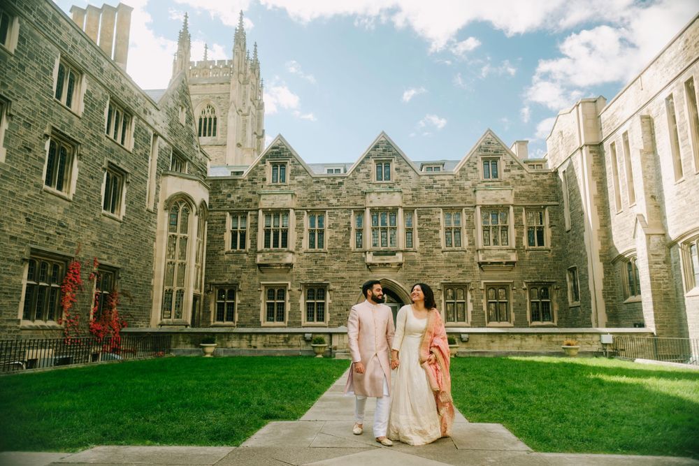 Three people in graduation attire walk across a courtyard of a Gothic style university building on a sunny day.