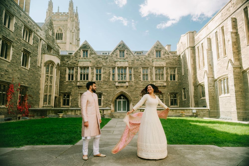 A couple poses on a manicured lawn in front of a grand Gothic Revival university building on a sunny day.