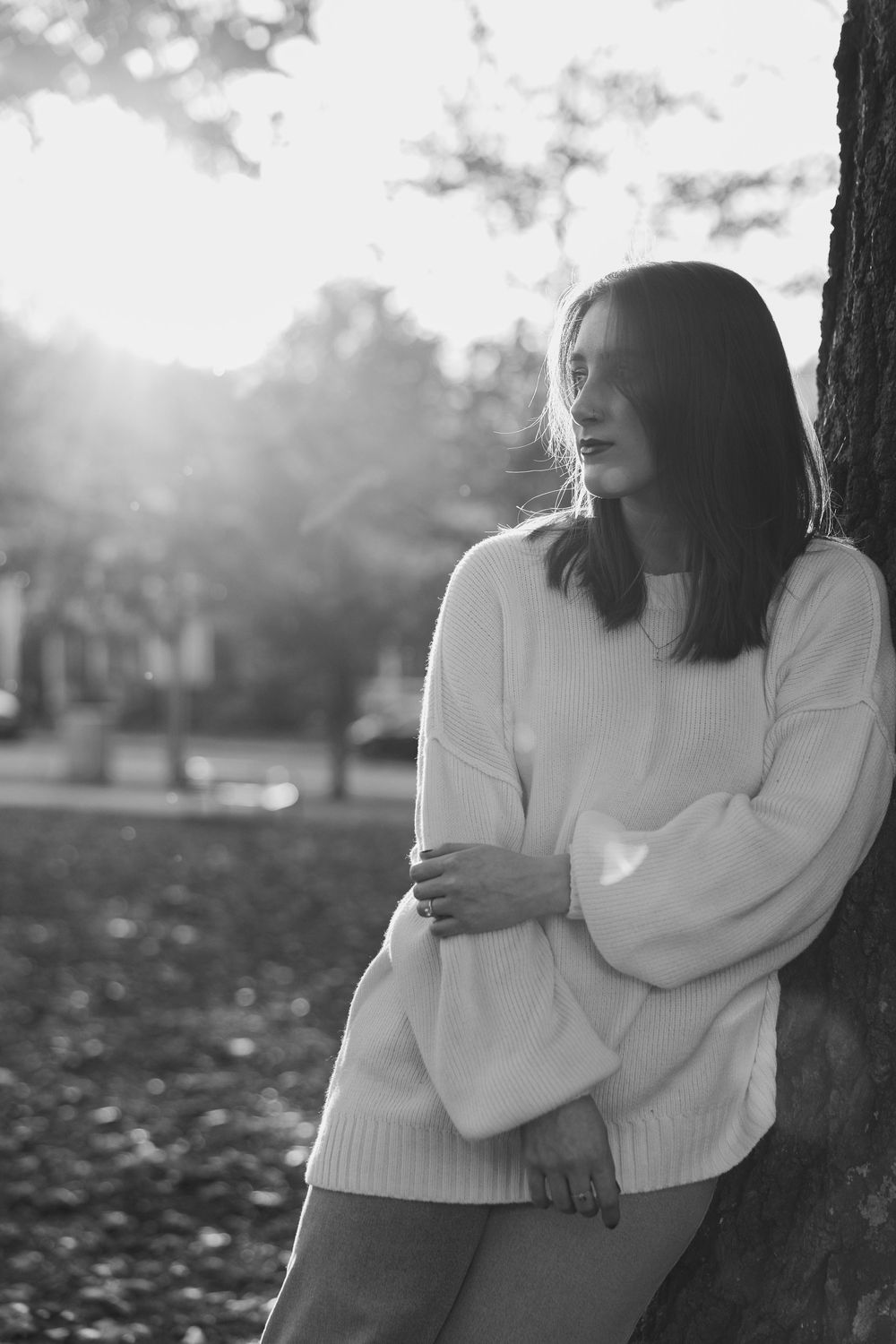 Black and white photo of a young woman in oversized sweater leaning against a tree in a sunlit park.