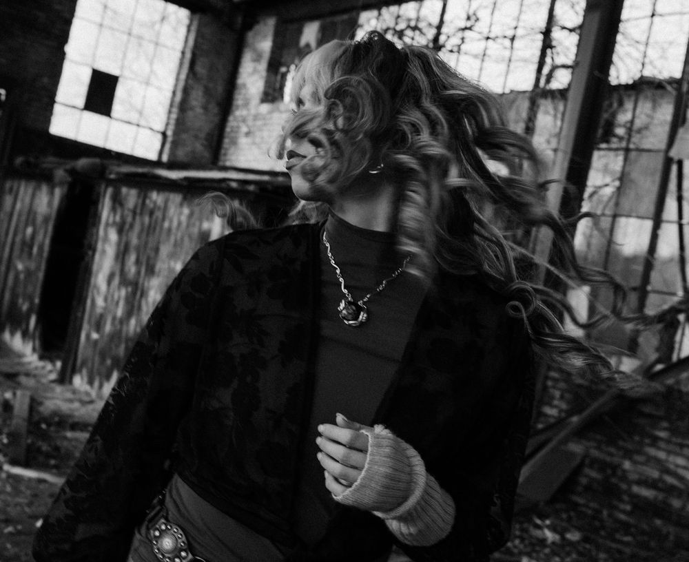 Black and white photo of a woman with flowing hair wearing a necklace, posed dramatically near an abandoned building.