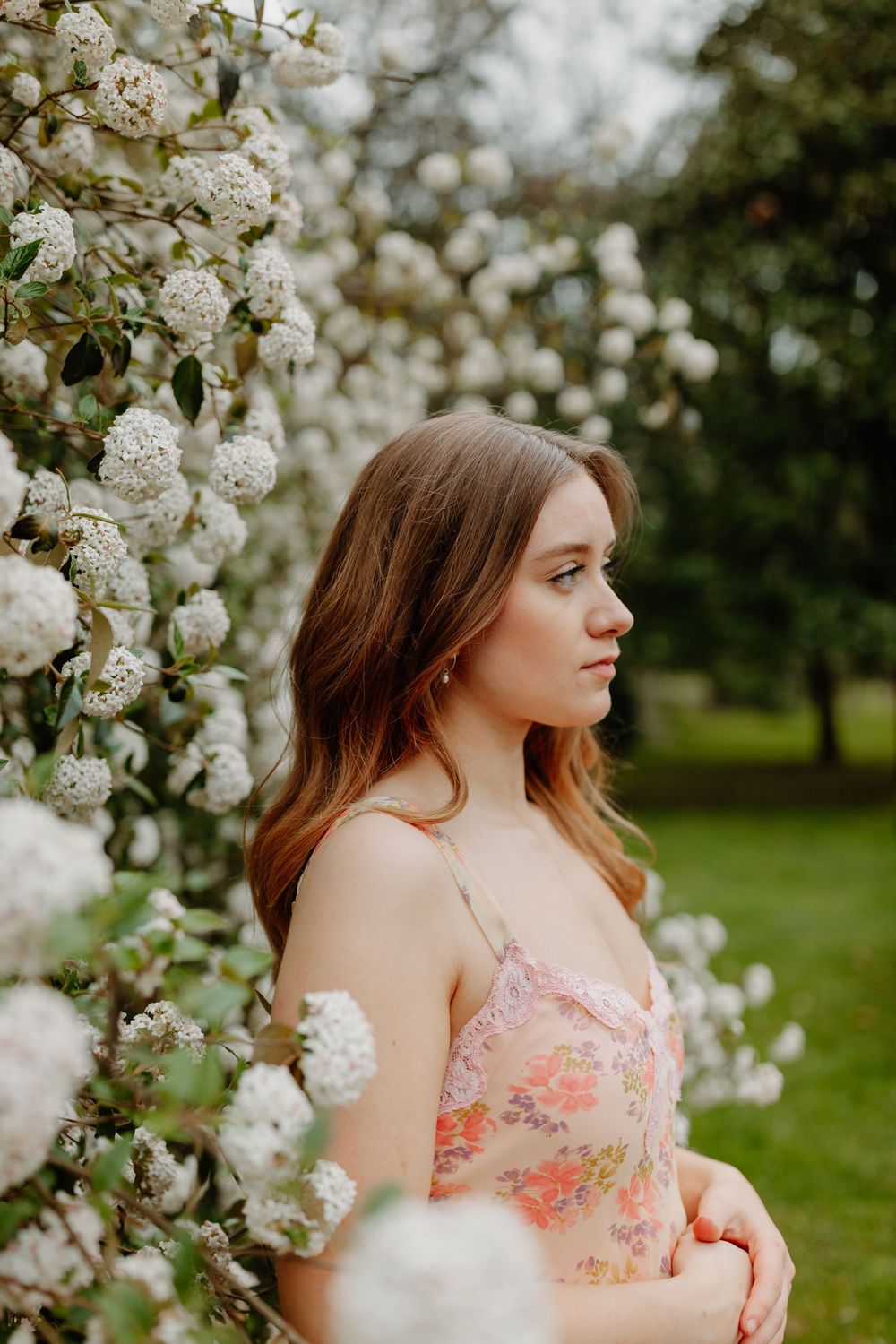 Young woman in floral dress standing beside blooming white flower bushes in a lush green garden.