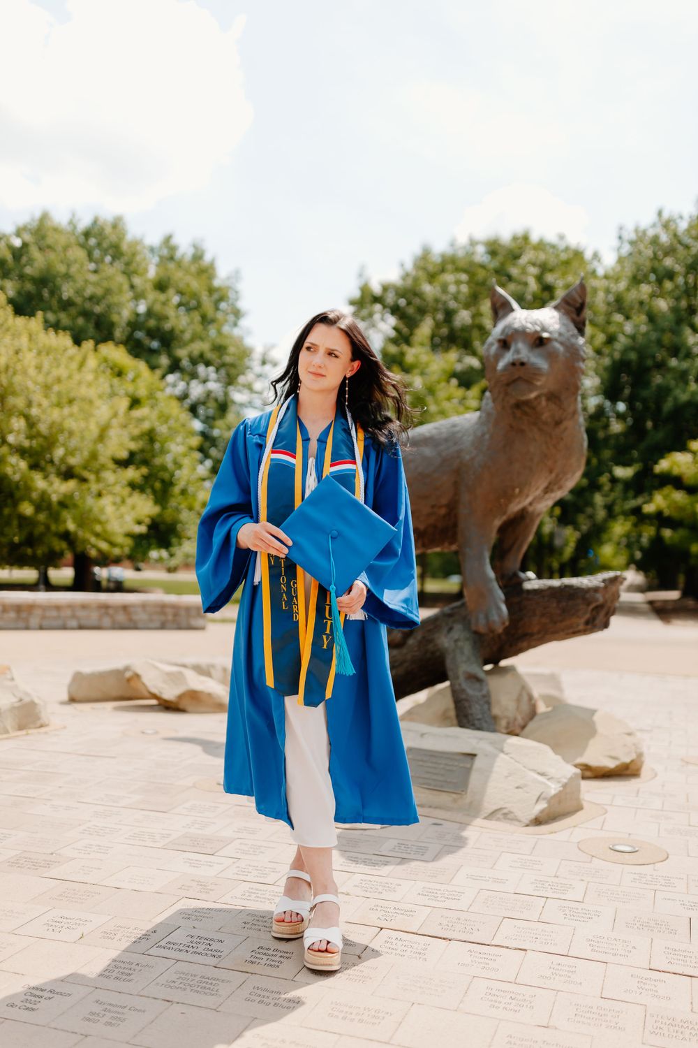 Graduate in blue cap and gown poses near university mascot statue for graduation photos.