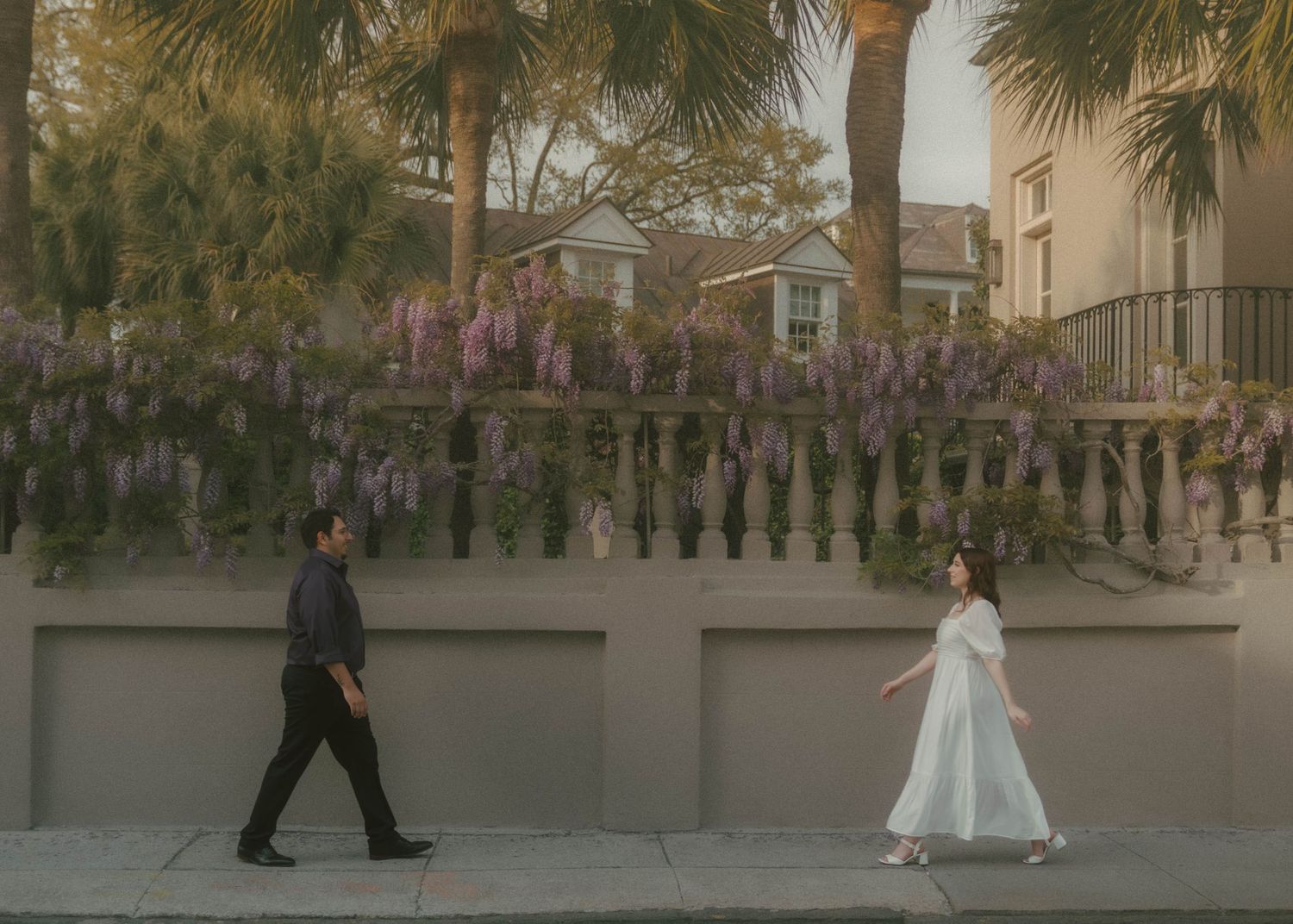 Two people walking in opposite directions on a sidewalk with wisteria flowers hanging from above and palm trees in background.