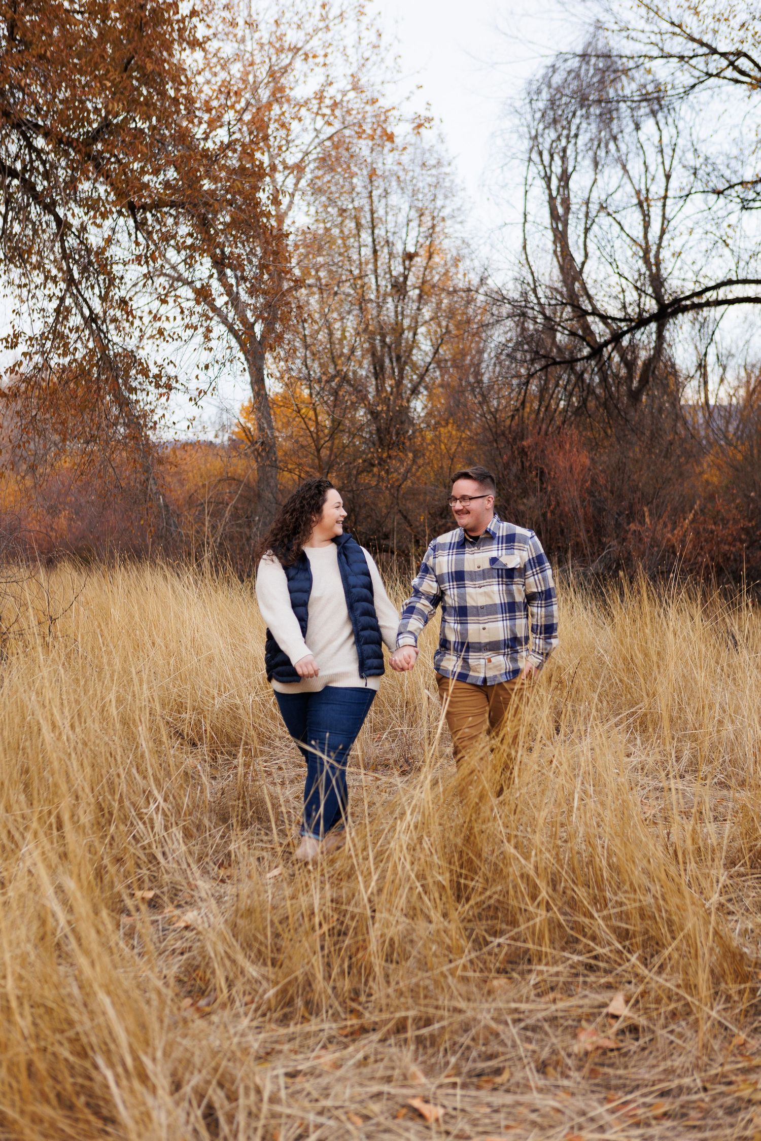 A couple walks together through tall autumn prairie grass surrounded by bare trees on an overcast day.