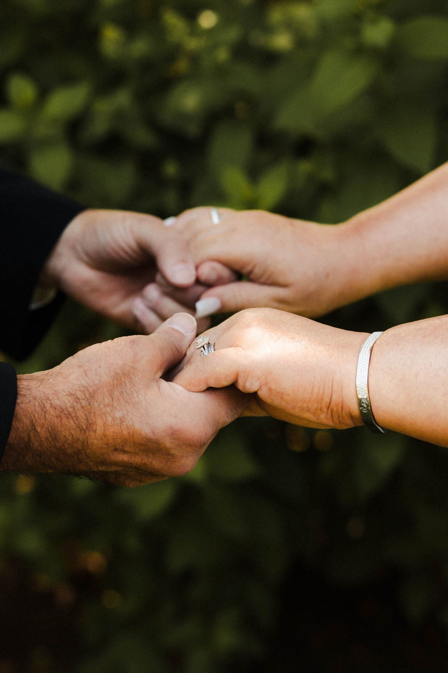 Two hands tenderly hold each other with a silver bracelet visible against a blurred green outdoor background.