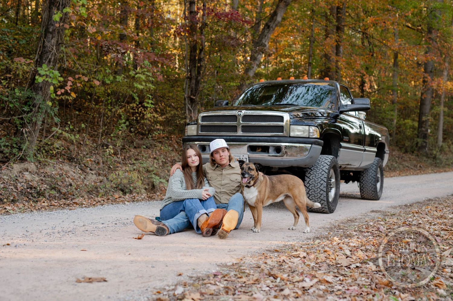 A dog sits loyally beside people relaxing near a lifted black pickup truck on a forest road.