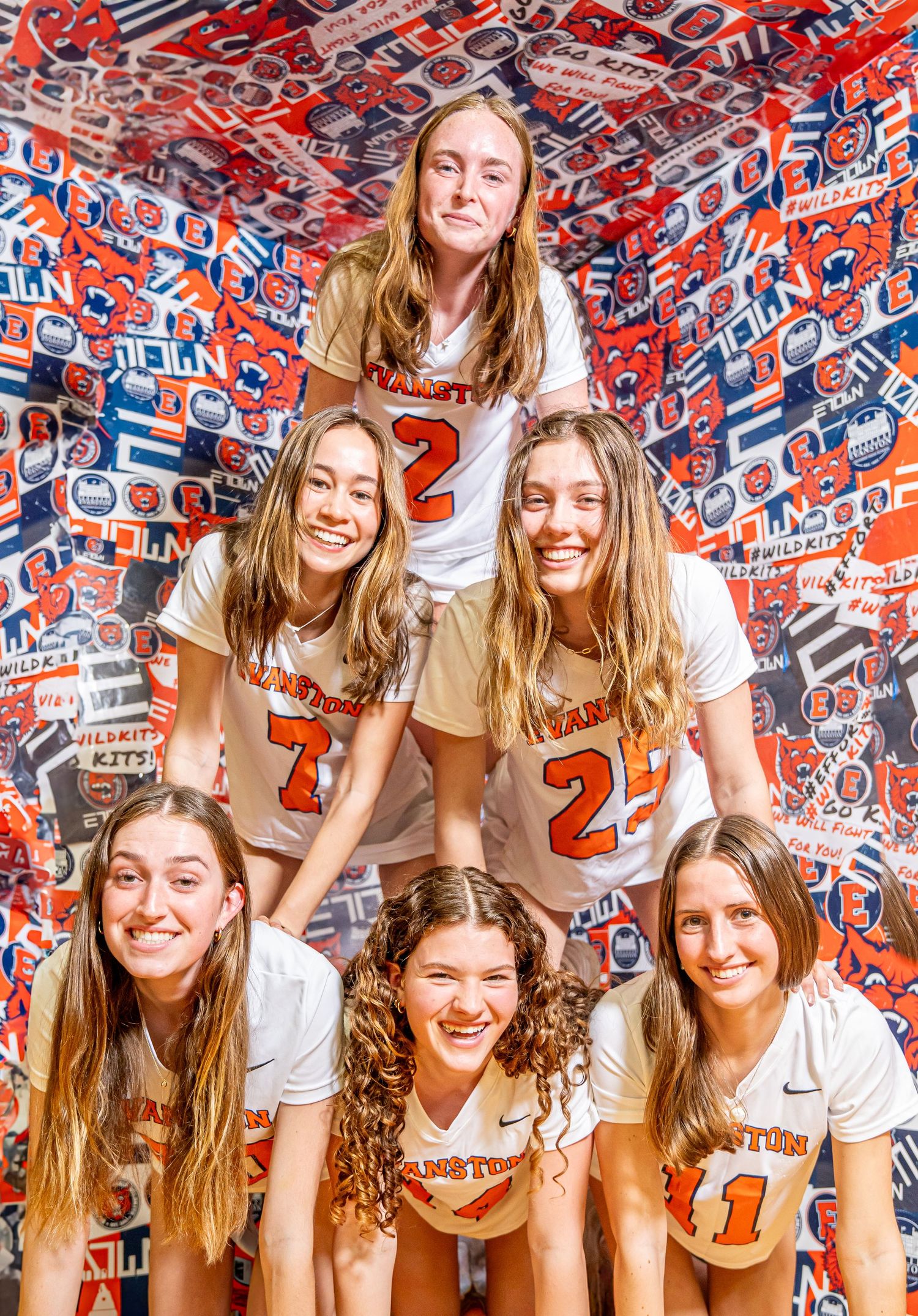 Group of athletes in white sports jerseys with numbers pose together against a red and blue patterned background.