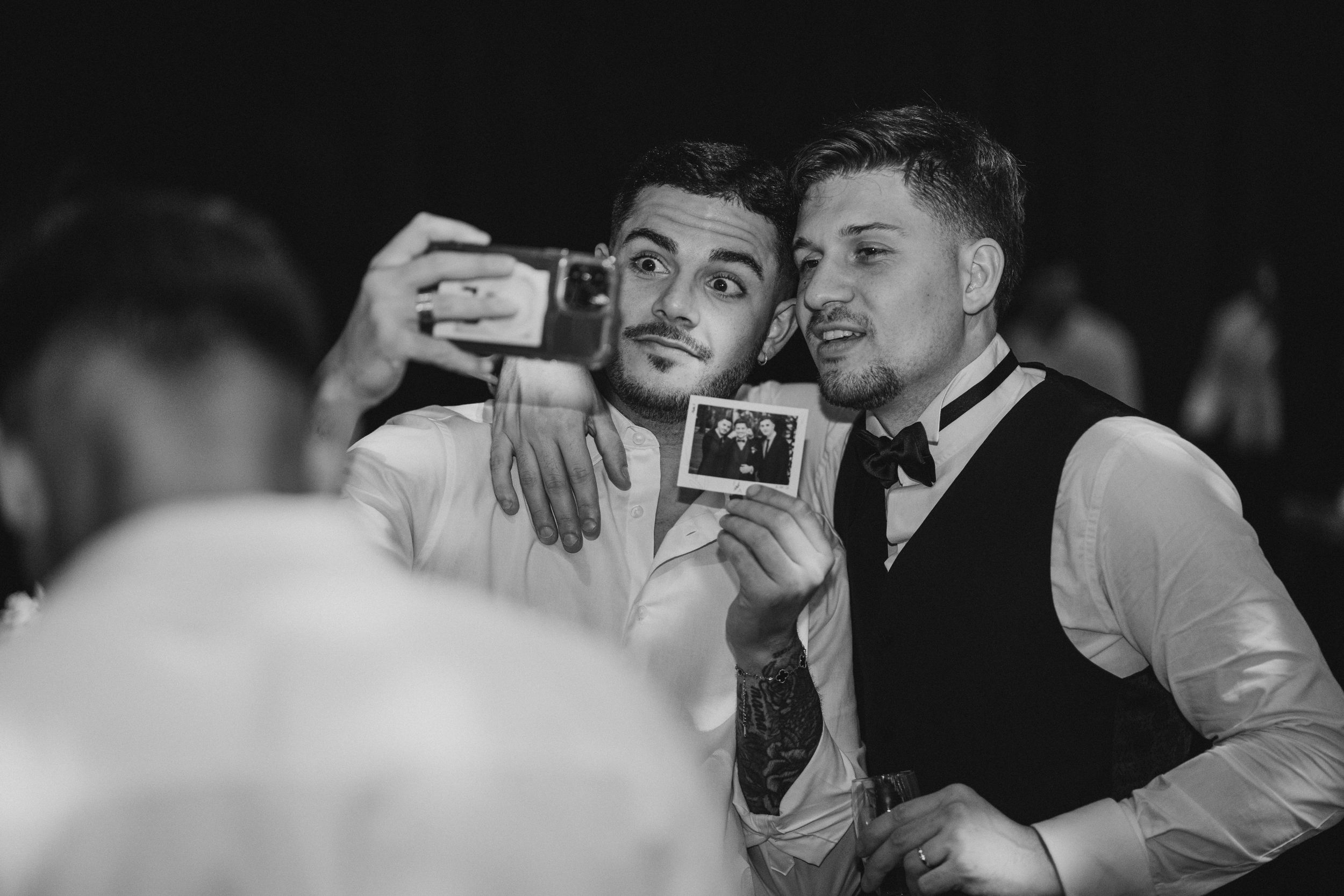 Two people in formal attire holding up a Polaroid photo and taking a selfie together at what appears to be an event.