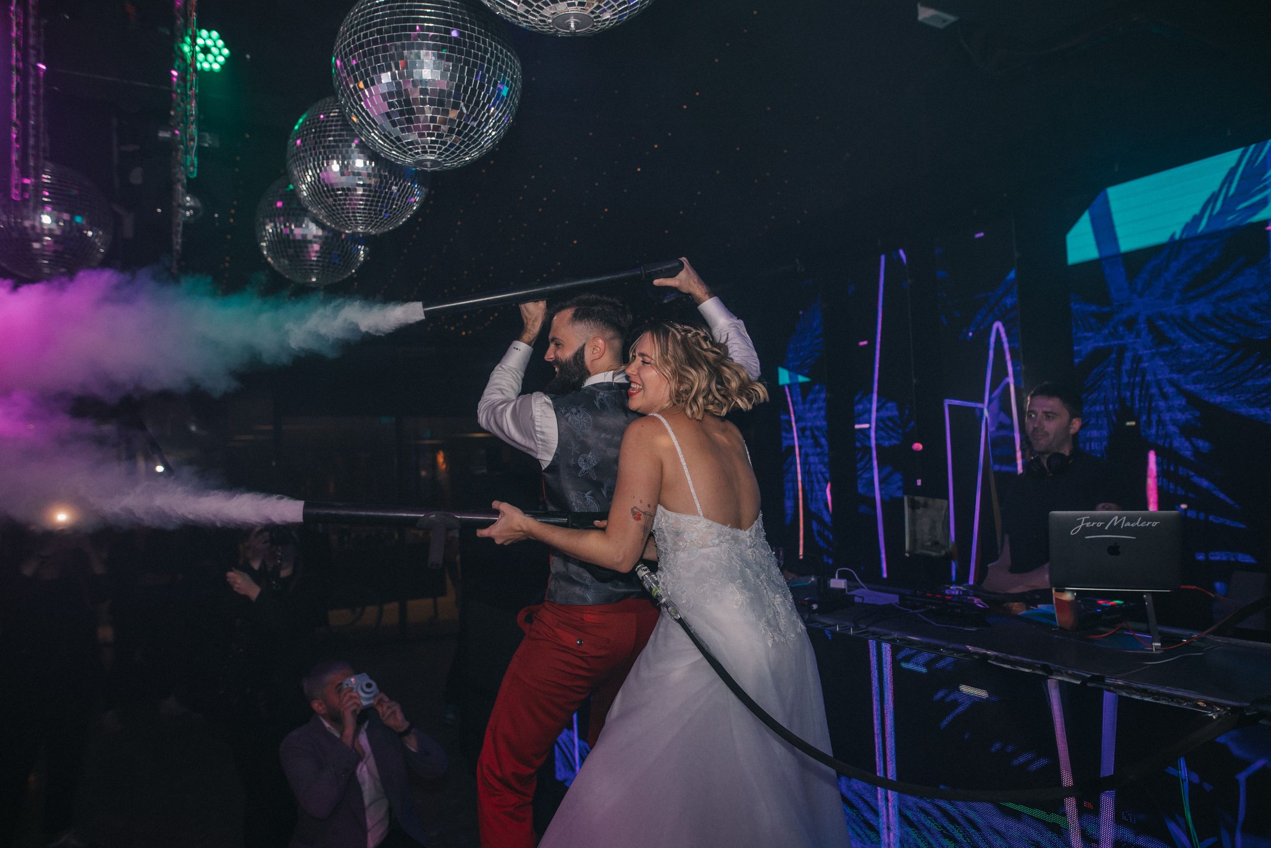 A couple dances under disco balls and purple lighting at a nightclub celebration with fog effects in the background.