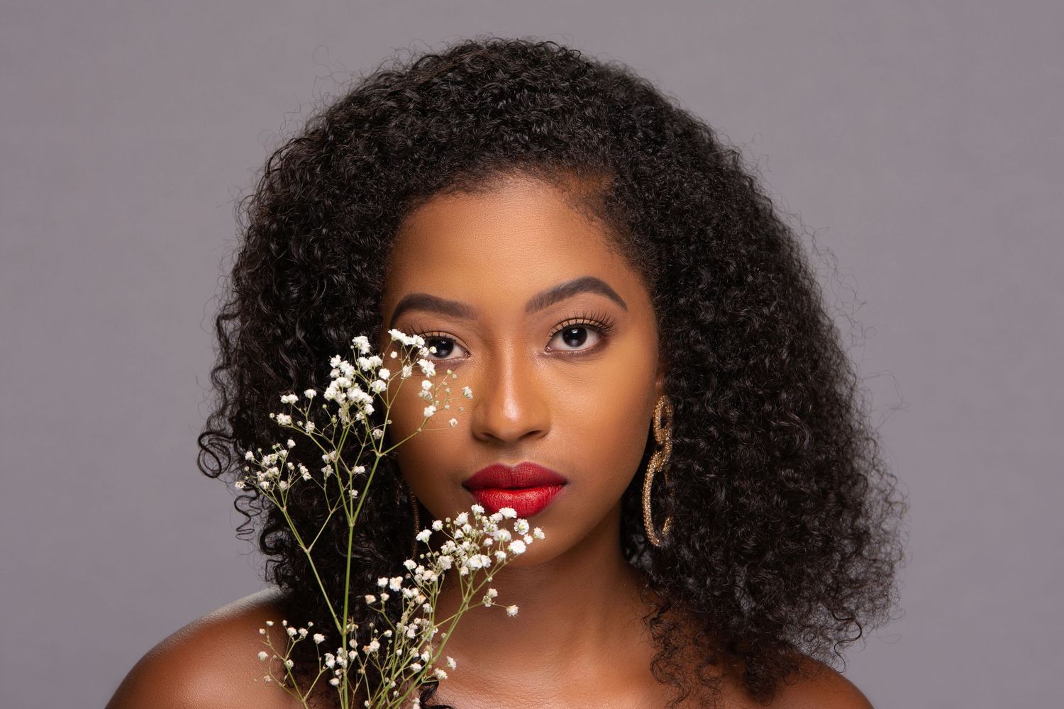 Young Black woman with curly hair holds white baby's breath flowers, wearing gold hoop earrings and red lipstick.