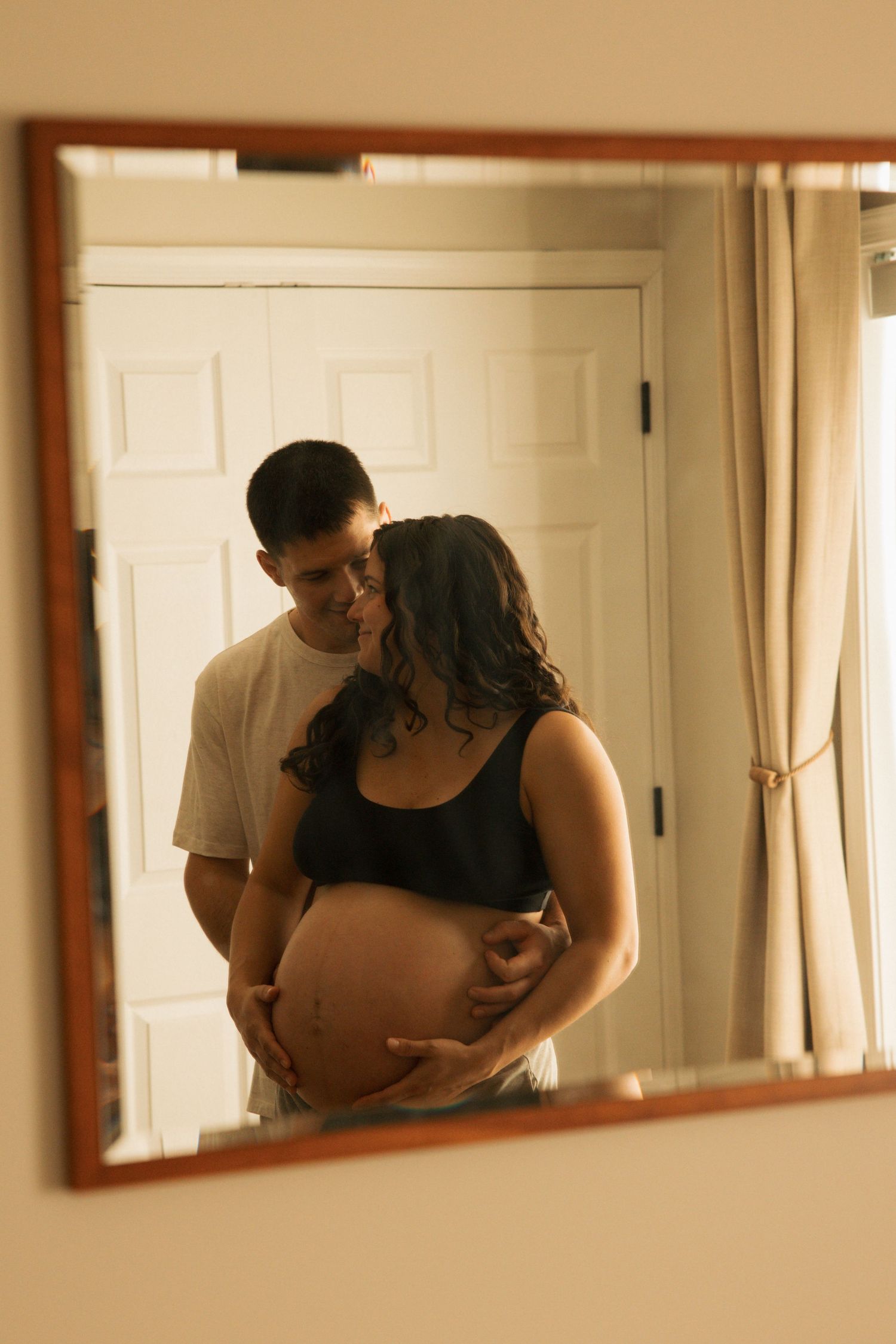 Couple embracing in mirror reflection, pregnant woman cradling baby bump with partner kissing her cheek in bedroom.