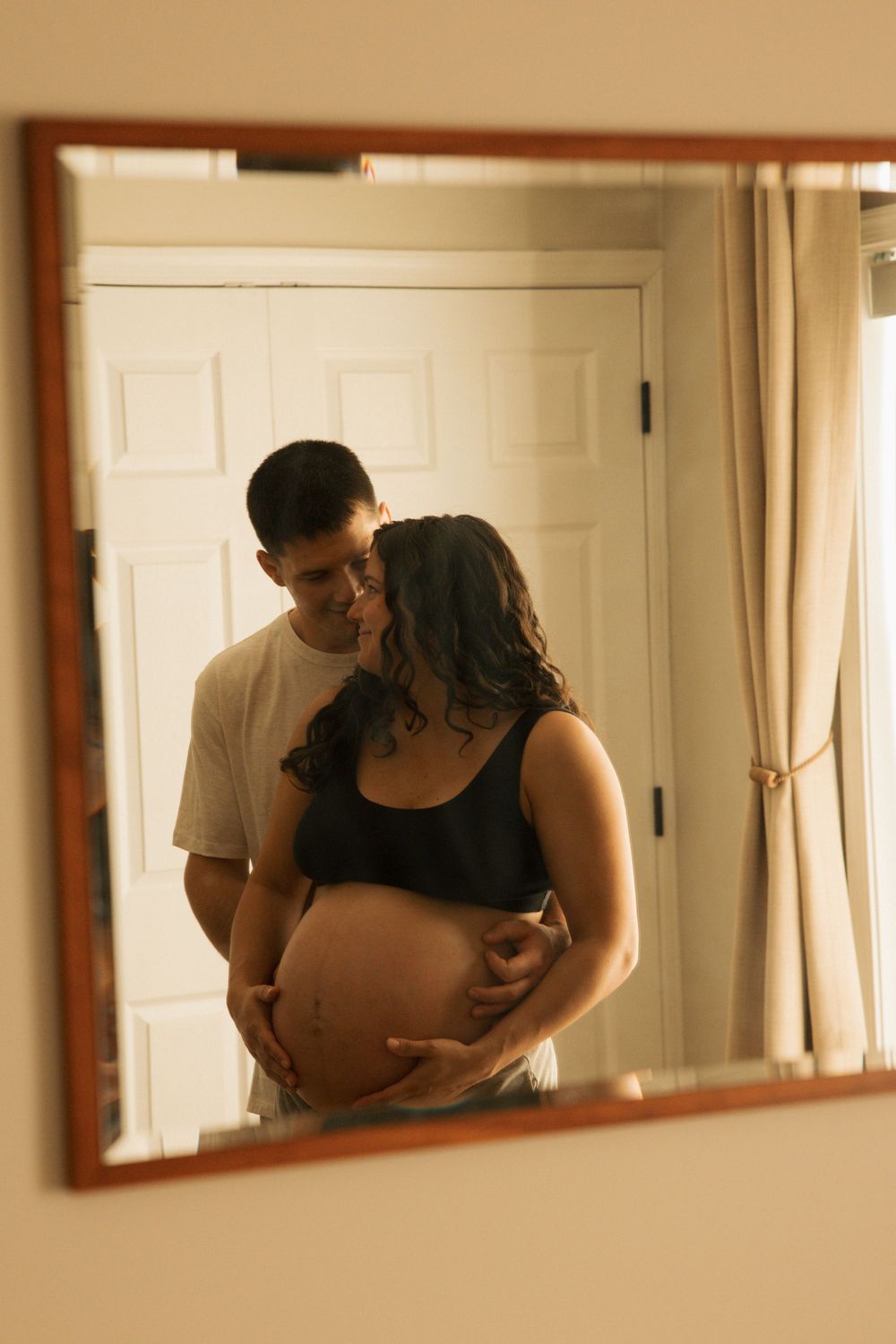 Couple embracing in mirror reflection, pregnant woman cradling baby bump with partner kissing her cheek in bedroom.