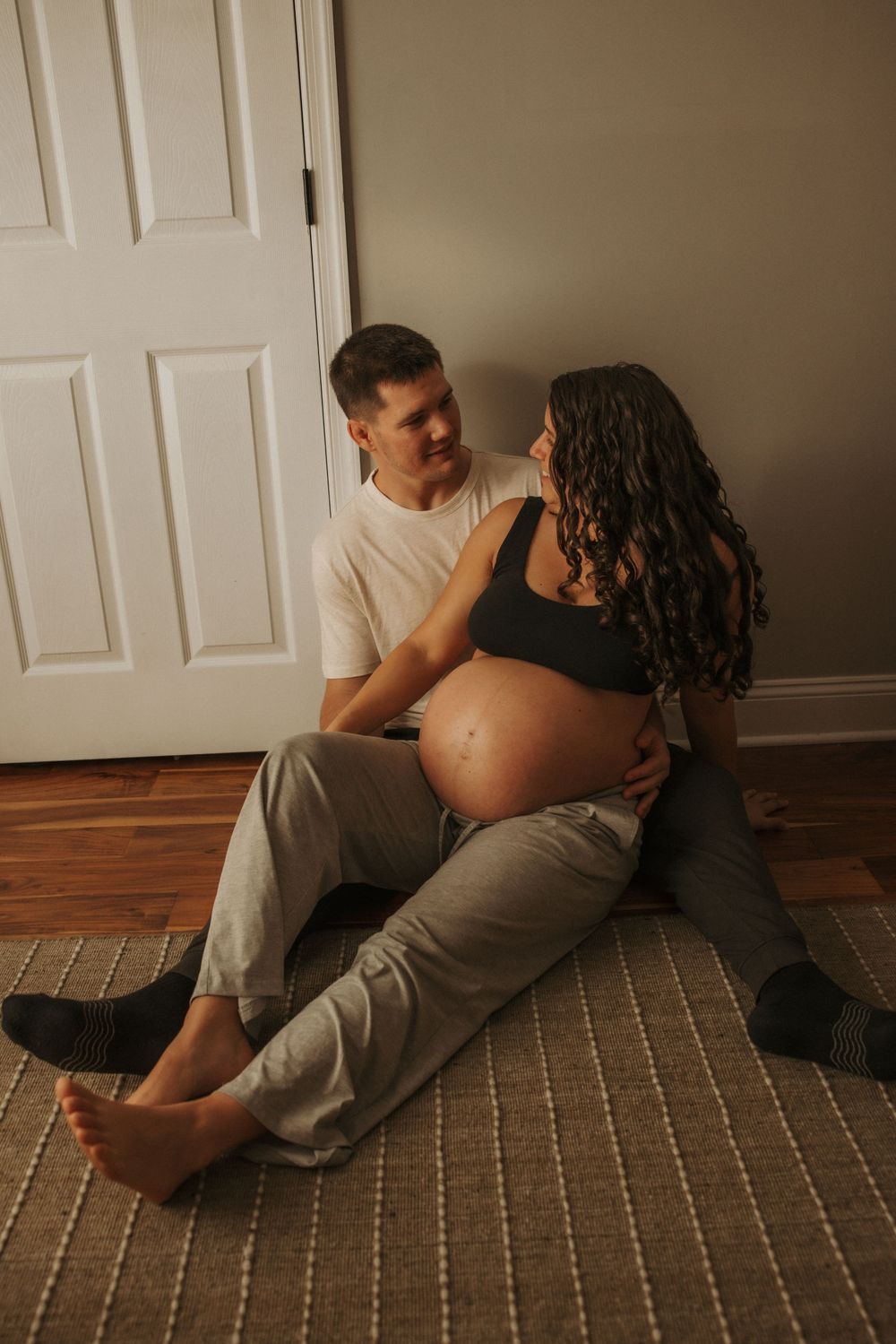 Couple sitting on floor, pregnant woman in black crop top with partner in white tee, sharing tender moment.