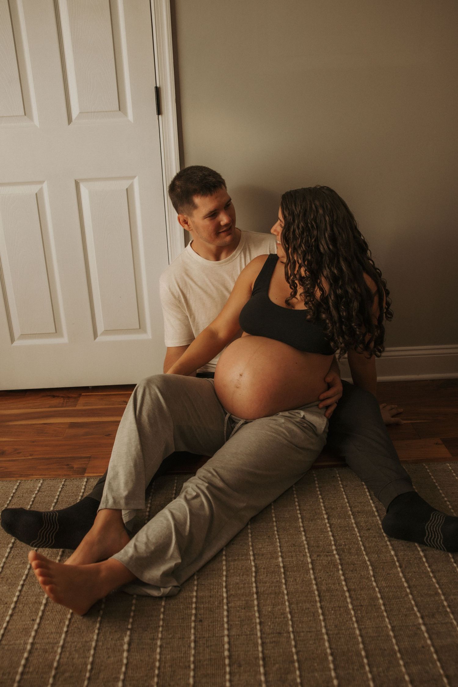 Couple sitting on floor, pregnant woman in black crop top with partner in white tee, sharing tender moment.
