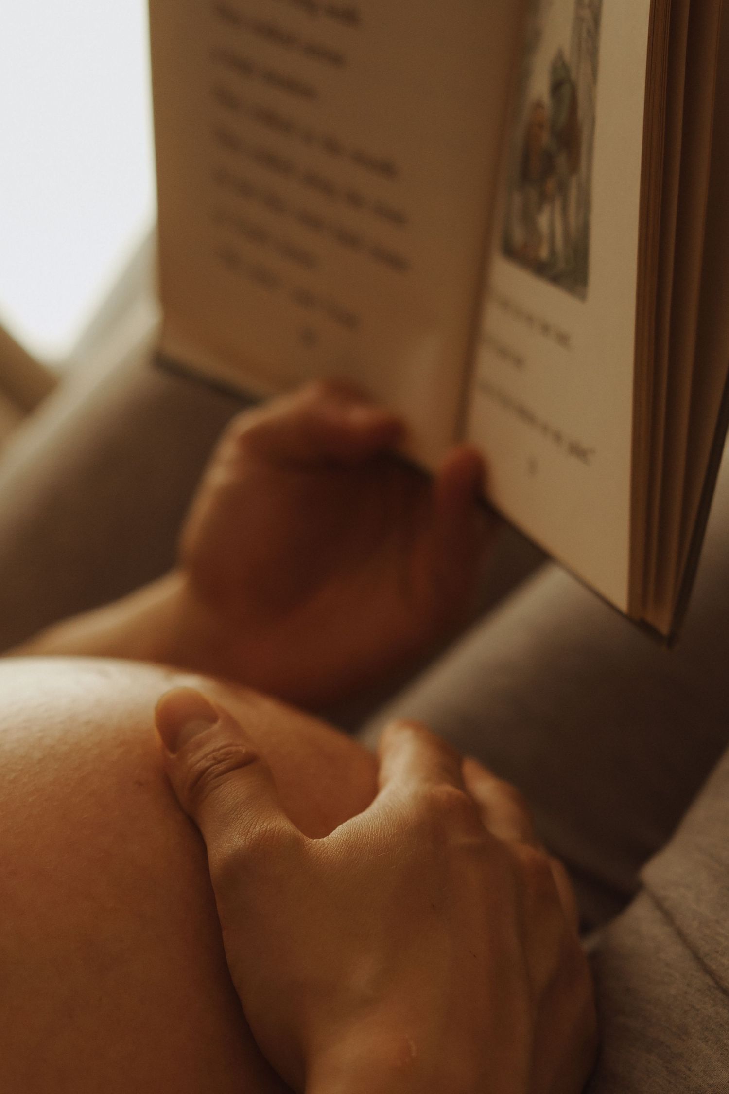Pregnant woman resting on a couch, holding her baby bump while reading a book in warm natural light.