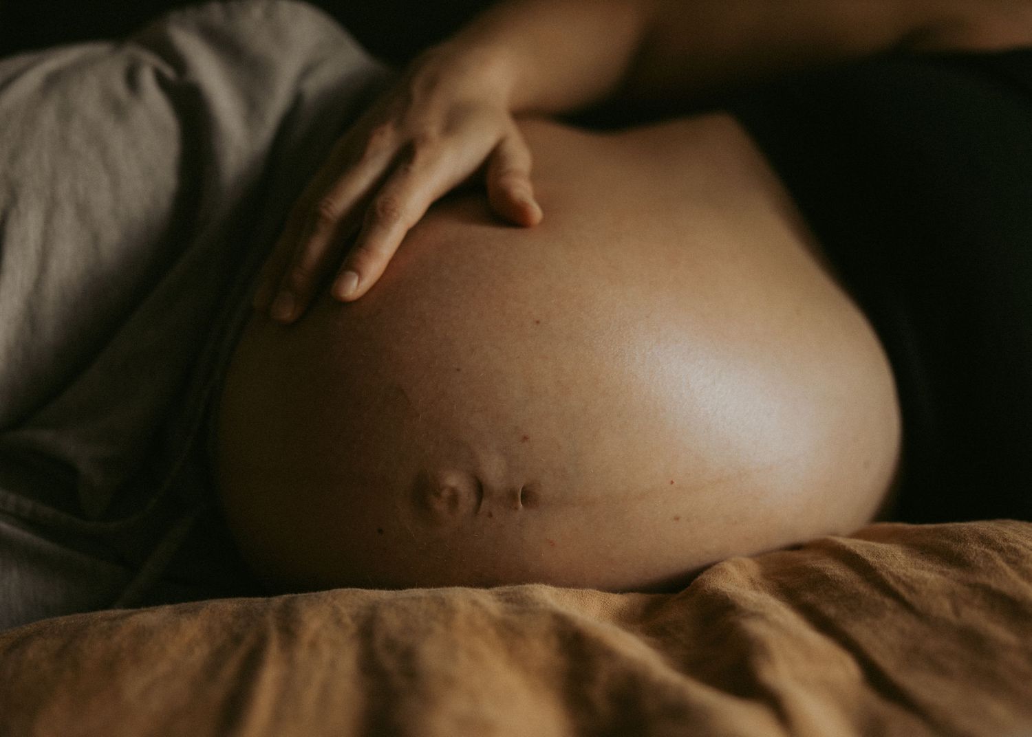 Close-up of a pregnant woman's bare baby bump as she rests on a bed with warm neutral tones.