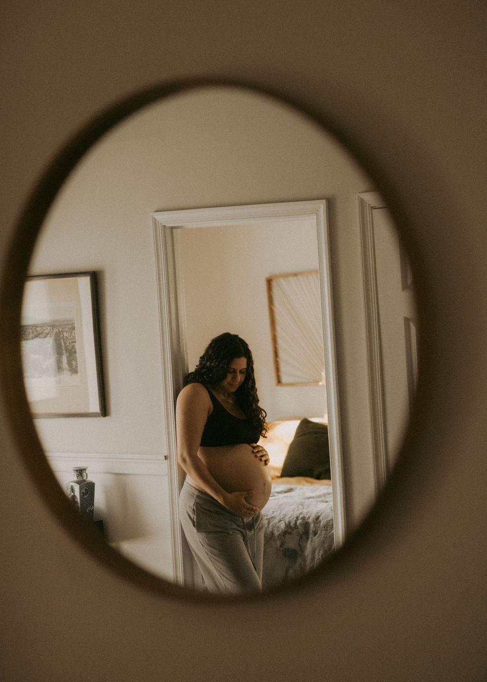 Pregnant woman cradles her baby bump, reflected in a round mirror in a warmly lit bedroom.