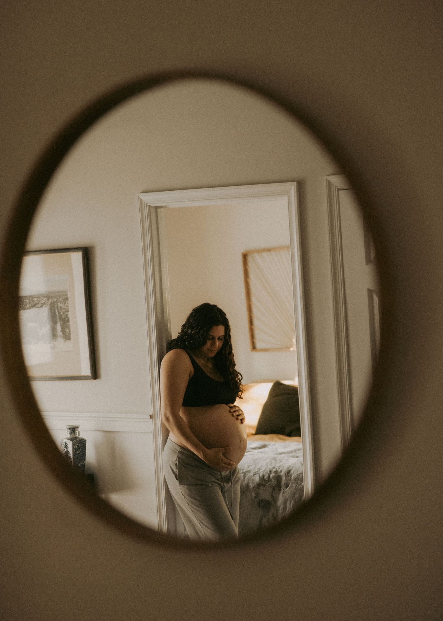 Pregnant woman cradles her baby bump, reflected in a round mirror in a warmly lit bedroom.
