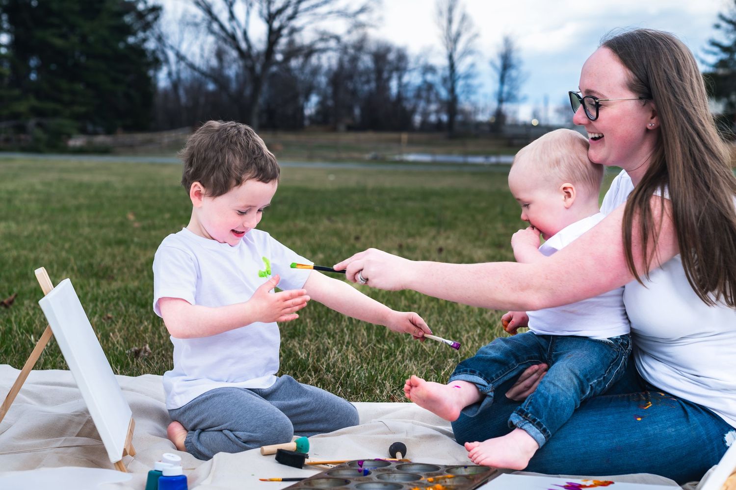 Family enjoying a sunny day outdoors with art supplies and a picnic blanket on the grass.