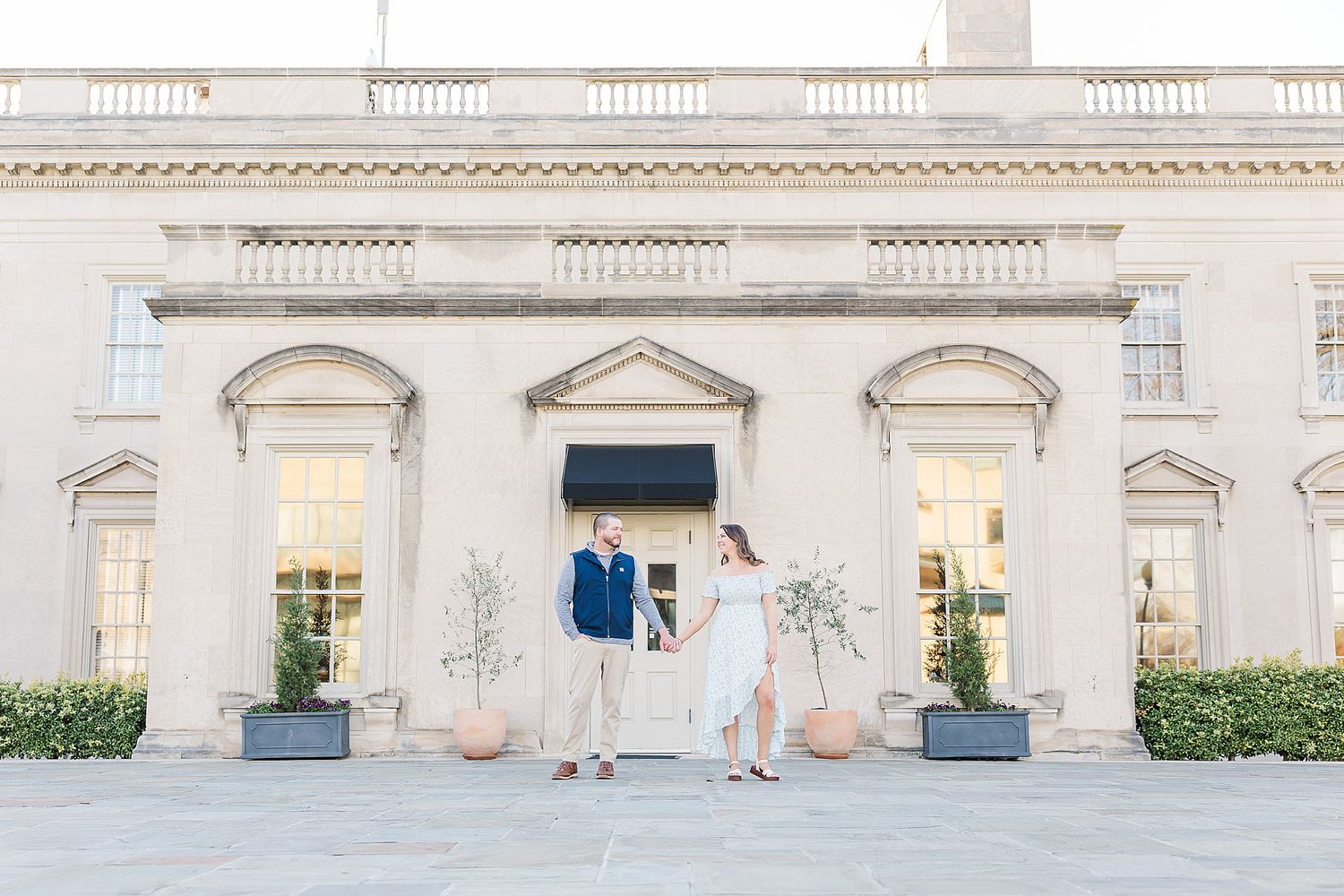 People stand outside an elegant historic mansion building with classical architecture and columns on a sunny day.