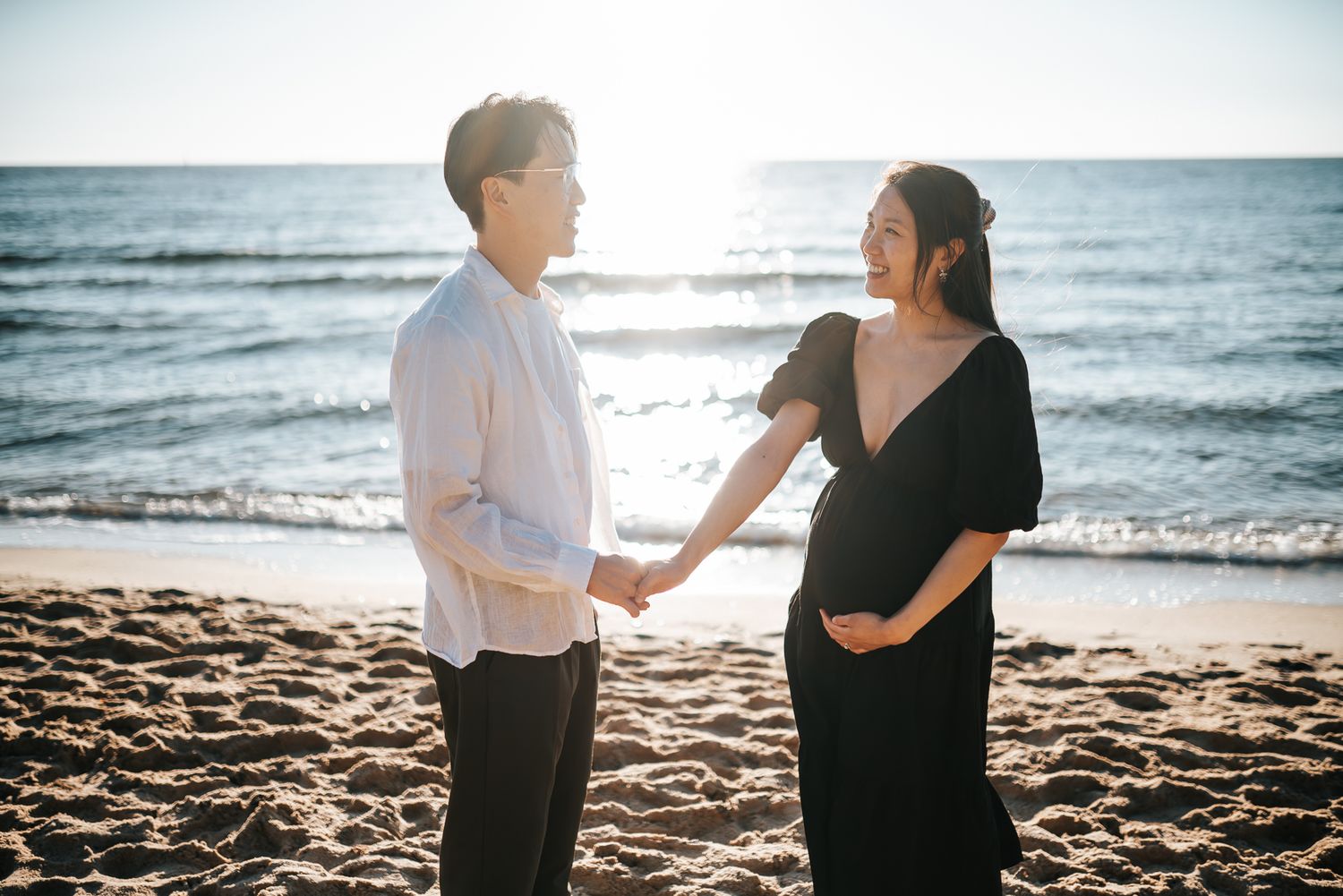 A pregnant couple walks hand in hand along a scenic beach at sunset during a Melbourne maternity photo session in Black Rock.