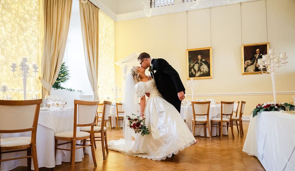 A couple shares a romantic dance moment in an elegant ballroom with golden curtains and wooden chairs.