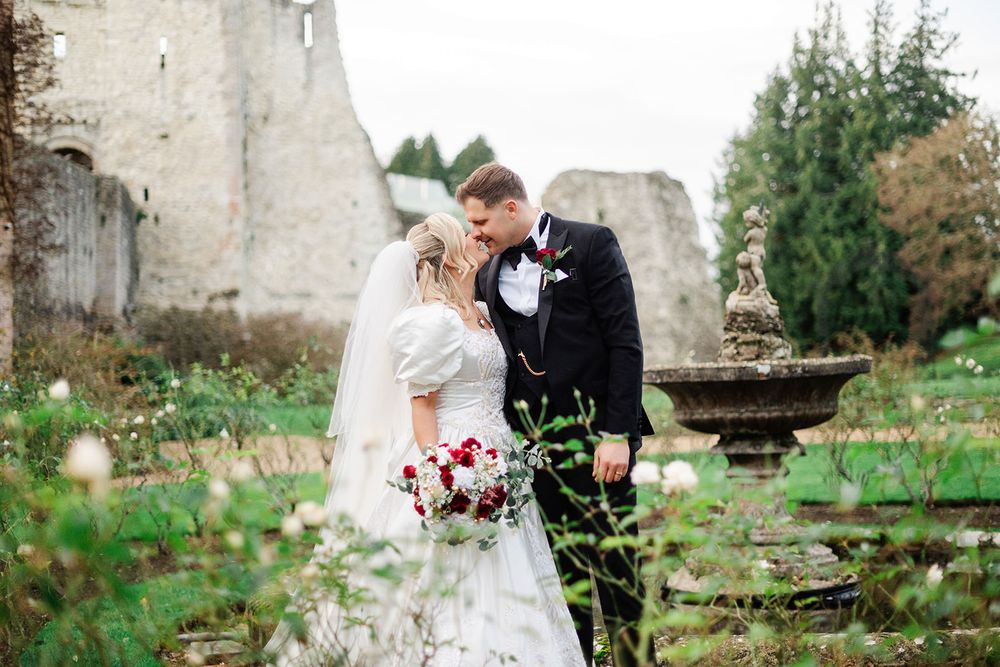 Newlyweds walk together through a lush garden setting with stone architecture and greenery in the background.