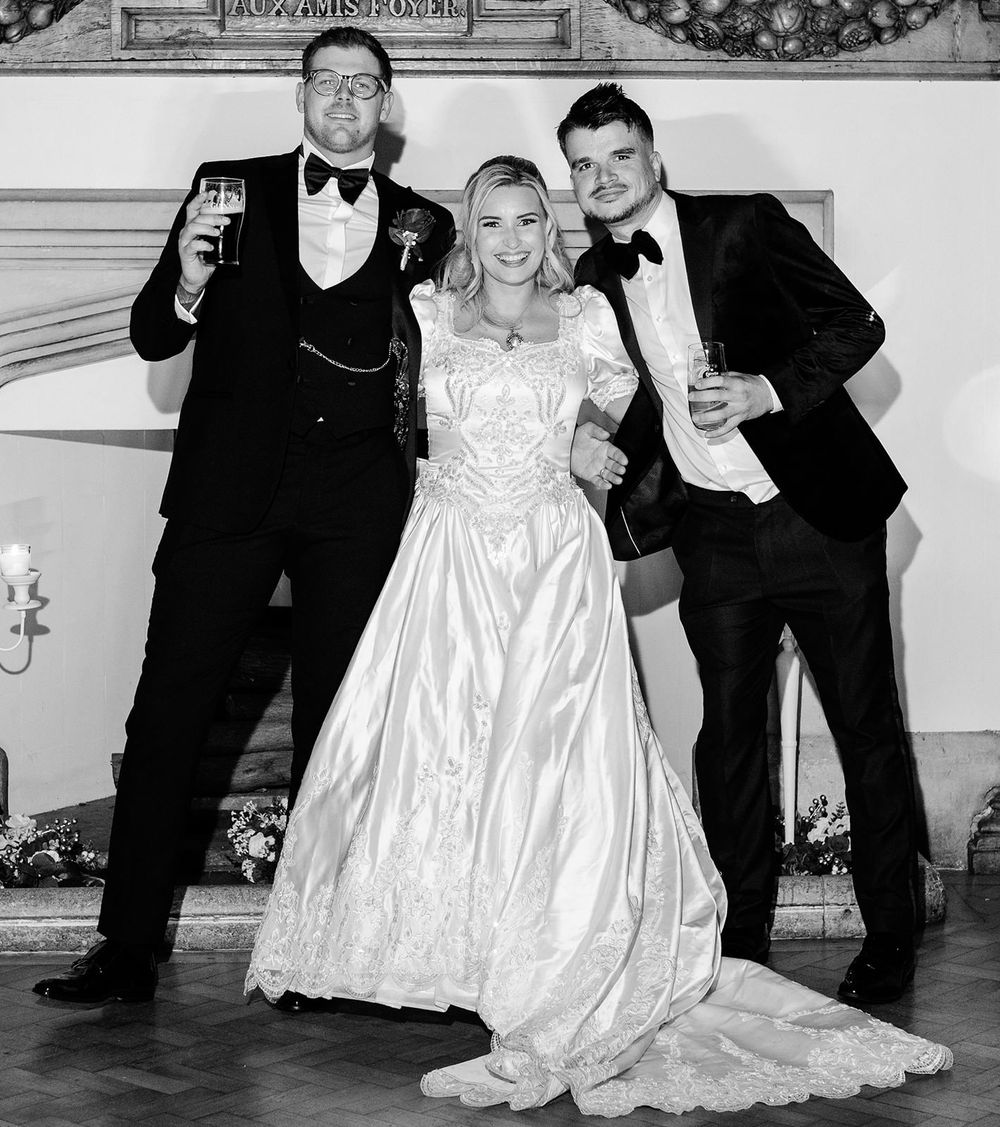 A black and white wedding photo showing a bride in a white gown posing with two people in tuxedos holding drinks.