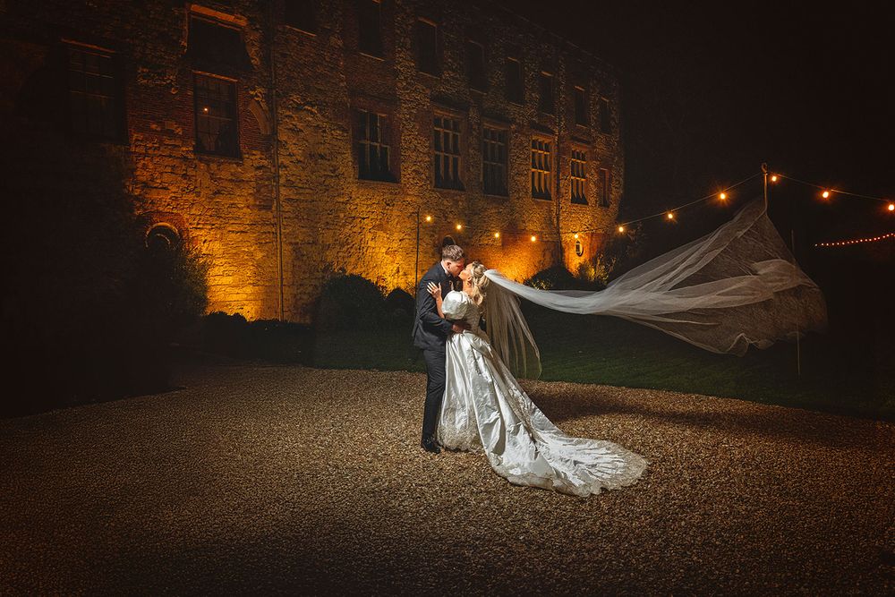 A romantic wedding couple poses at night in front of a beautifully lit stone castle with dramatic lighting.