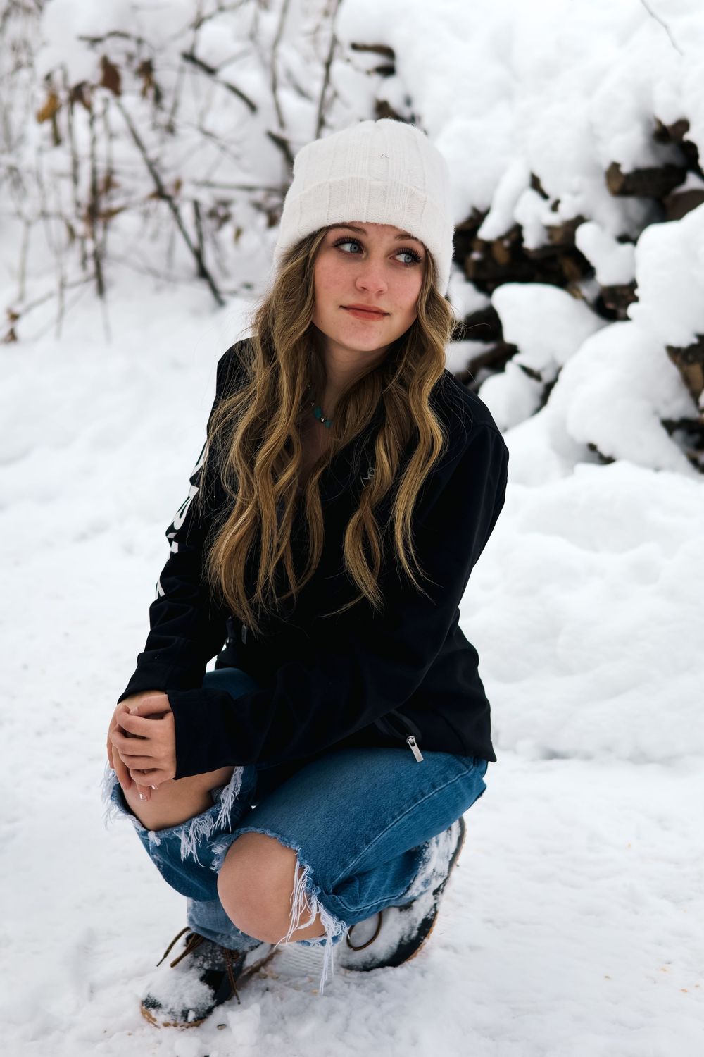 Teen girl in white beanie and ripped jeans crouches in snowy winter outdoor setting.