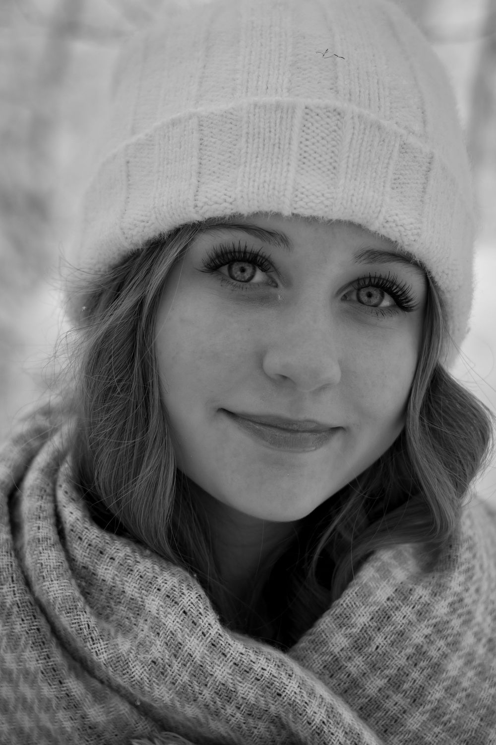 Black and white portrait of young woman in white knit hat and scarf in winter setting.