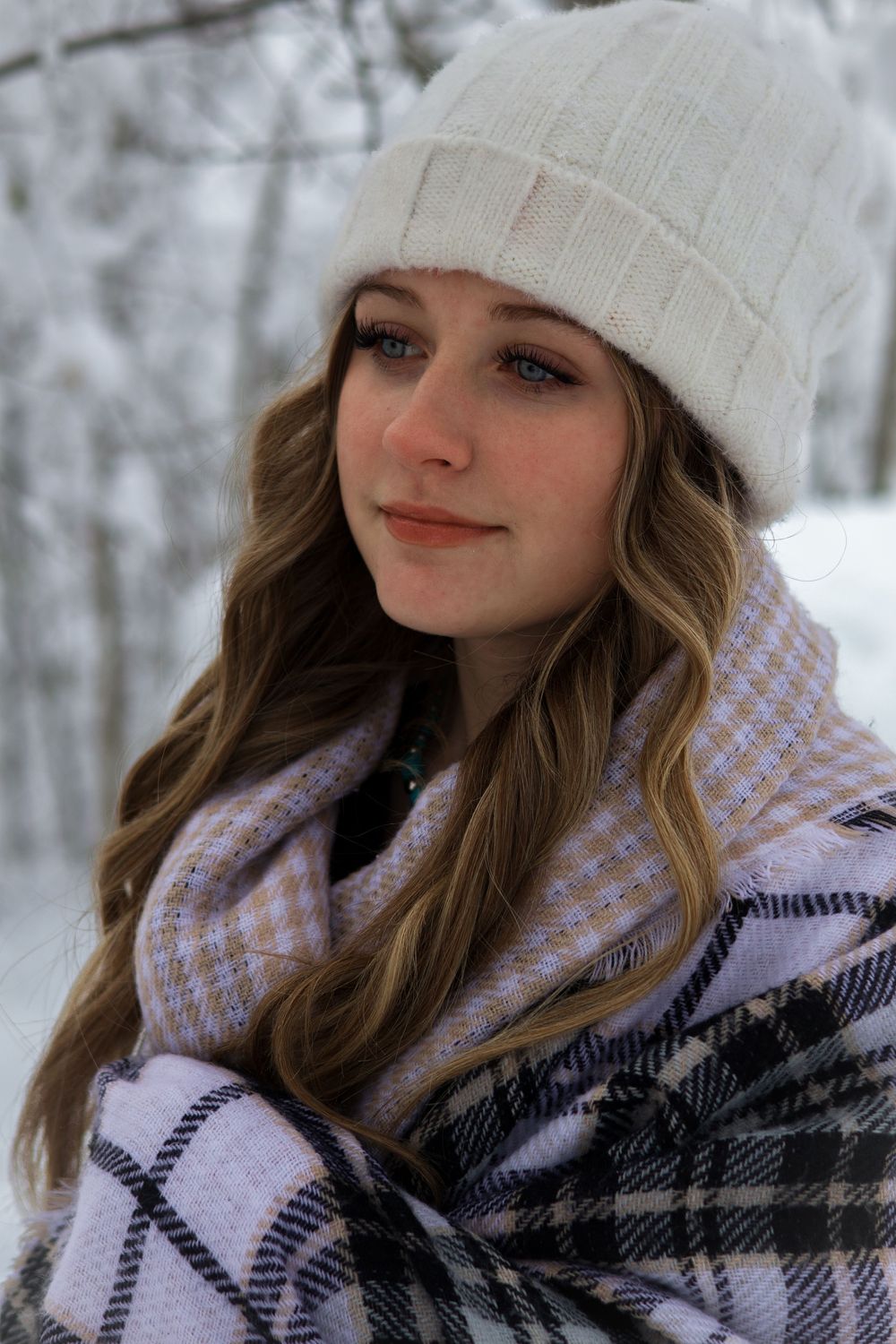 Young woman wrapped in plaid blanket scarf wearing white knit hat in snowy winter scene.