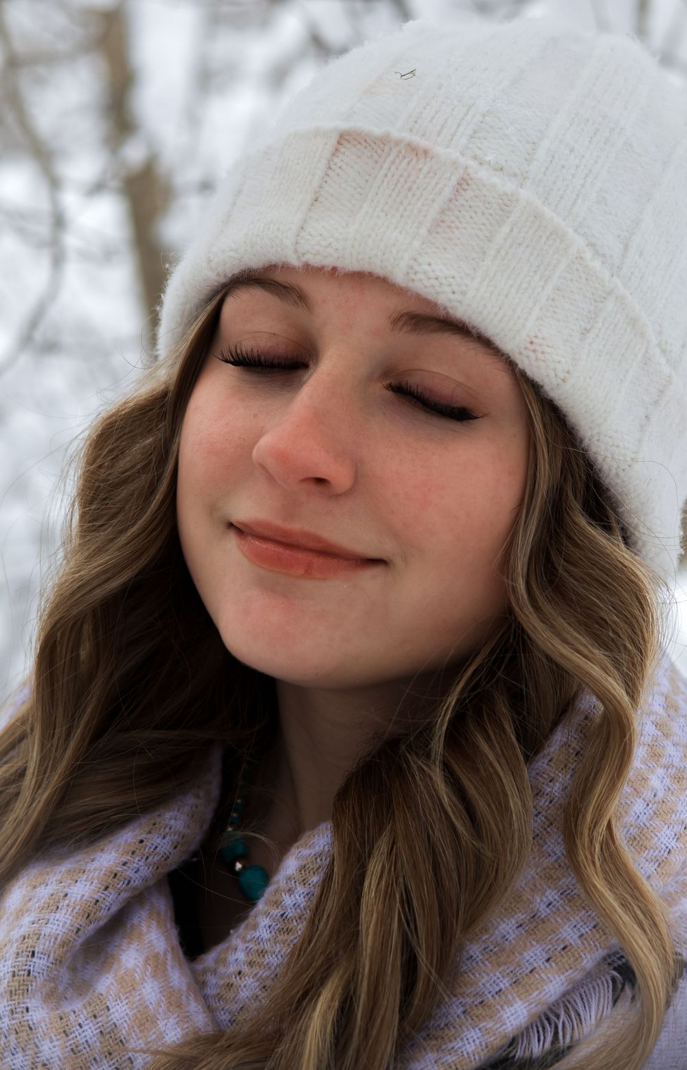 Young woman in white beanie smiles serenely with eyes closed in snowy winter landscape.