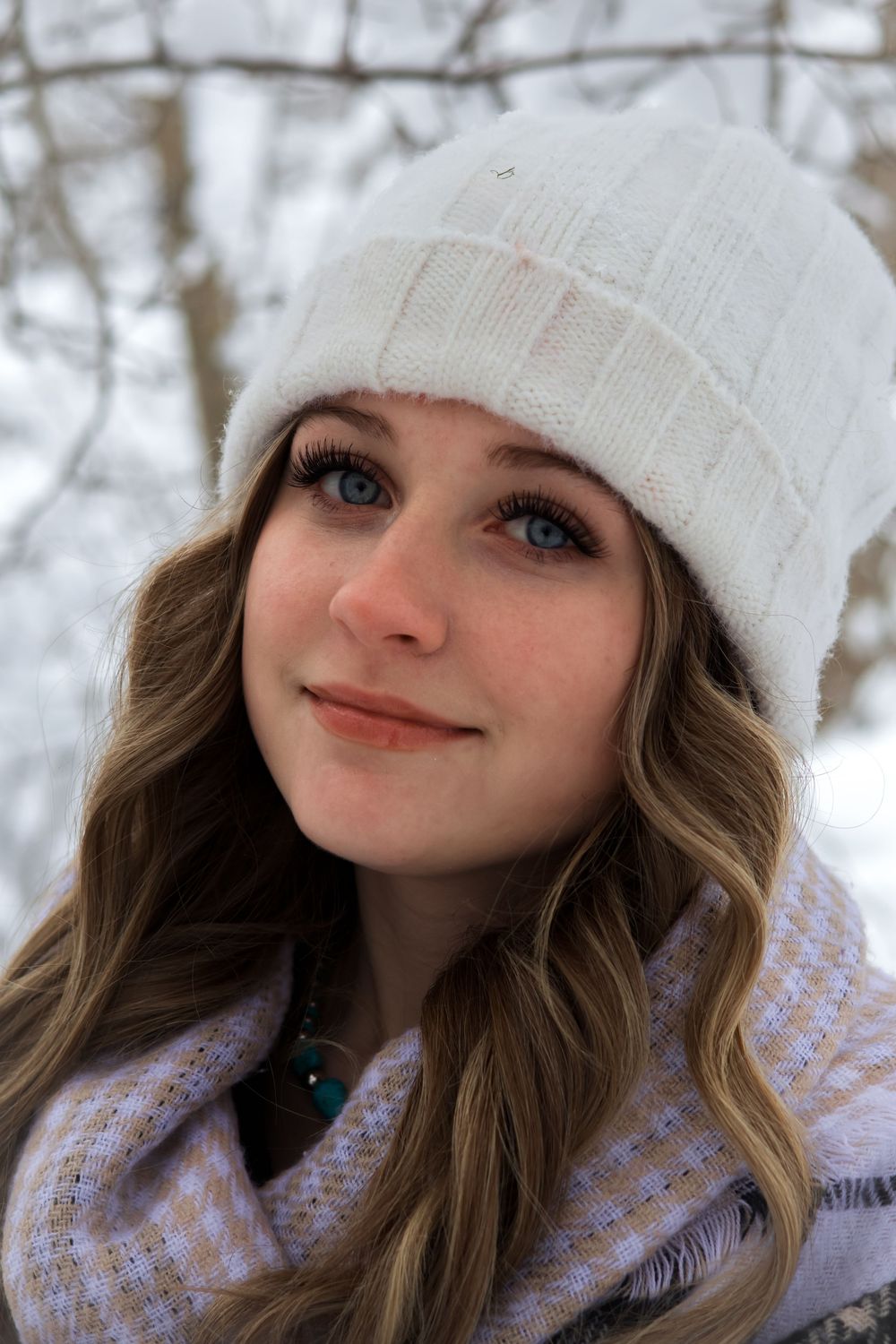 Blue-eyed young woman in white knit beanie and scarf smiles in snowy winter landscape.