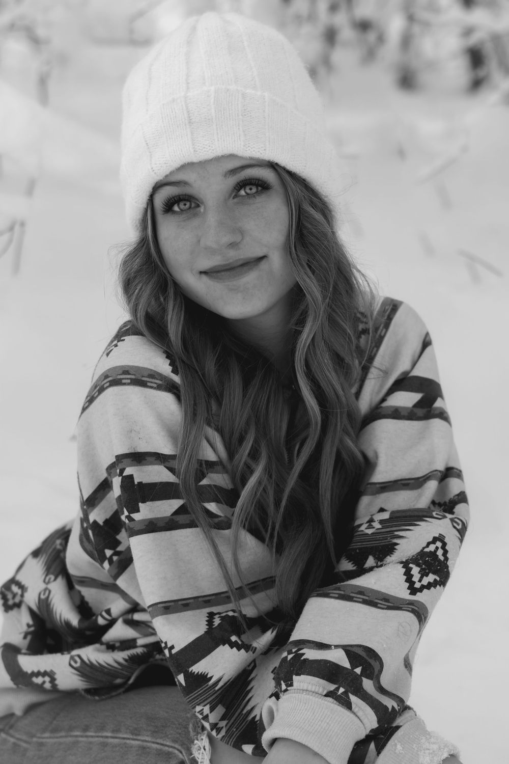 Black and white portrait of smiling teen girl in white beanie and patterned sweater in snow.